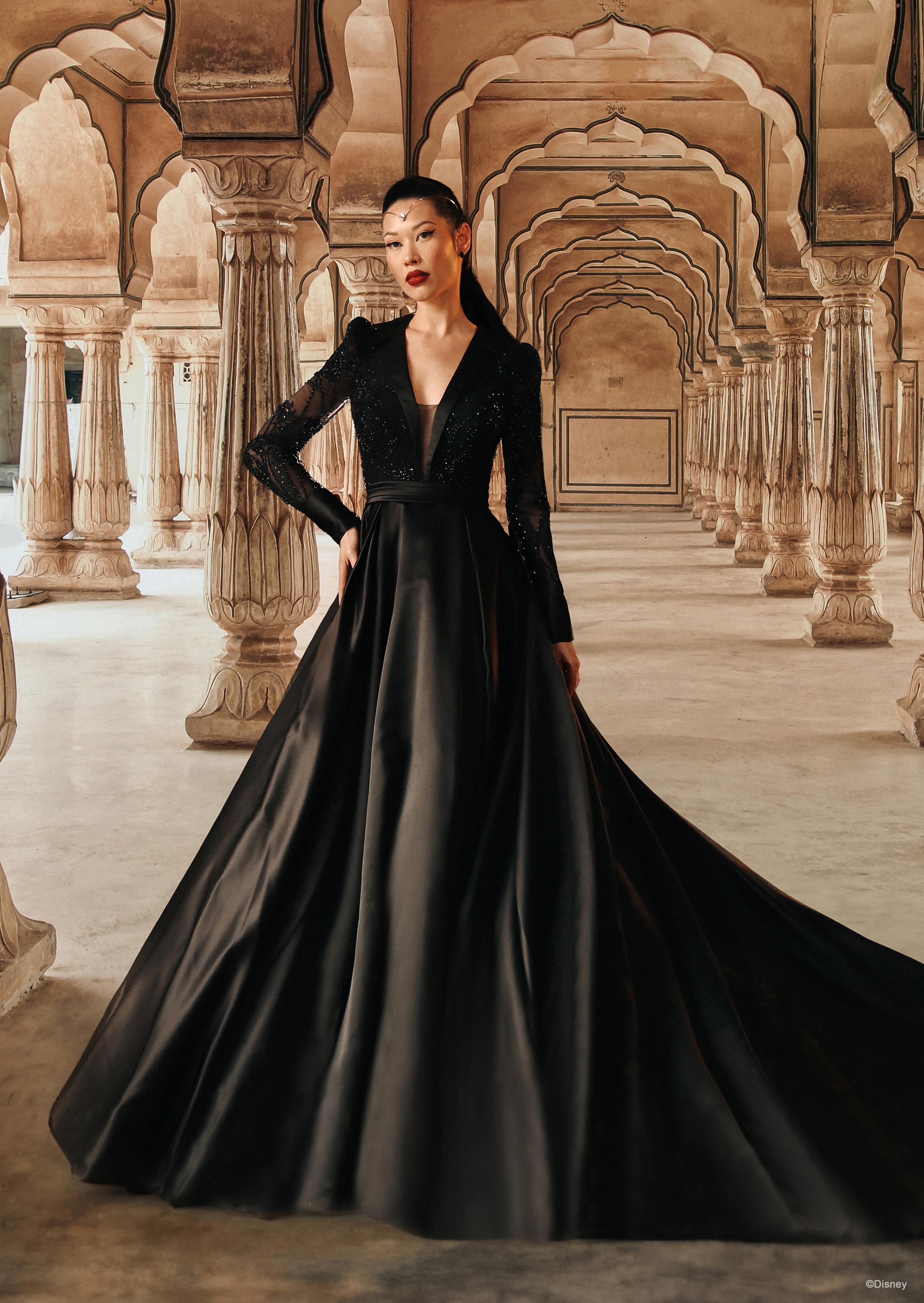 The image shows a woman wearing an elegant and sophisticated black wedding gown. The dress features a deep V-neckline and long sleeves, creating a sleek and refined look. The bodice is fitted, accentuating her waist, and the skirt is full and voluminous, flowing gracefully to the floor and extending into a dramatic train behind her. The fabric appears rich and luxurious, adding to the overall grandeur of the gown. The woman's hair is styled in a sleek updo, complementing the elegant and sophisticated design of the dress. The overall effect is one of timeless elegance and dramatic beauty, perfect for a non-traditional and striking wedding.