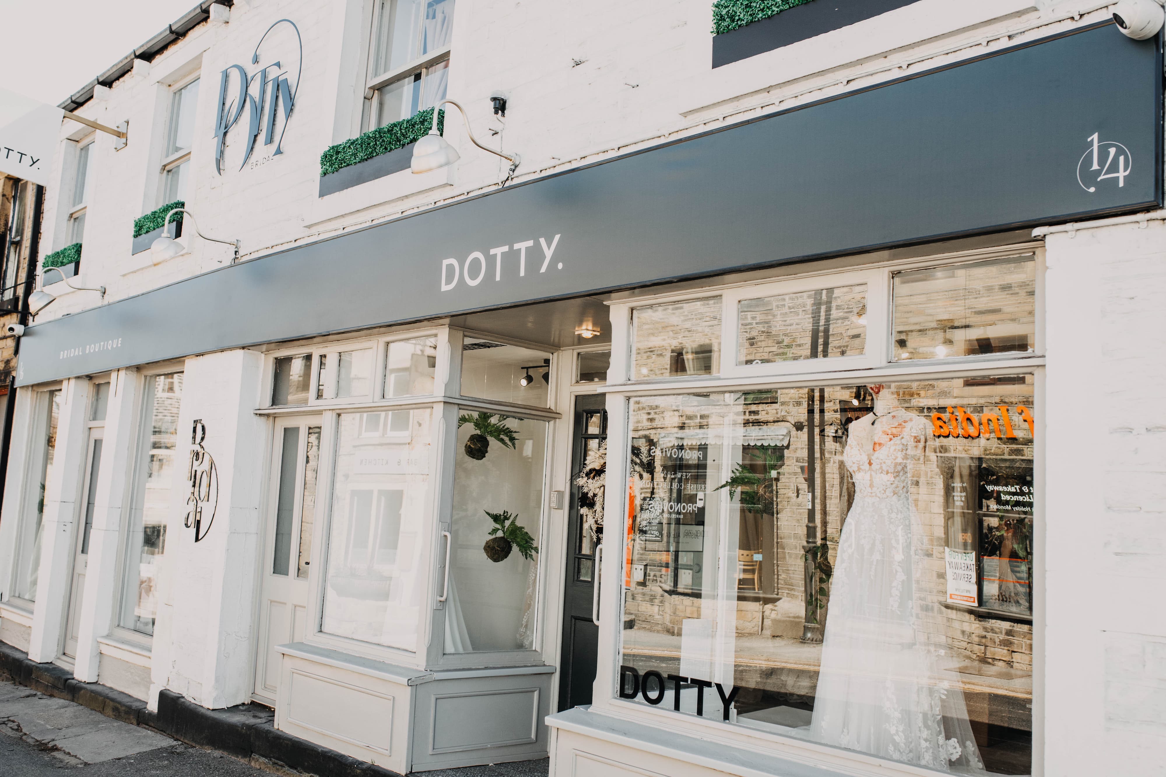 Exterior view of Dotty Bridal Boutique, showcasing its modern, minimalist design. The shop features large windows displaying elegant wedding dresses. The storefront is painted in light tones with a sleek, grey awning bearing the boutique’s name, "DOTTY," in clean, white lettering. The entrance is inviting with glass doors and potted greenery hanging inside, creating a chic and welcoming atmosphere for brides-to-be.