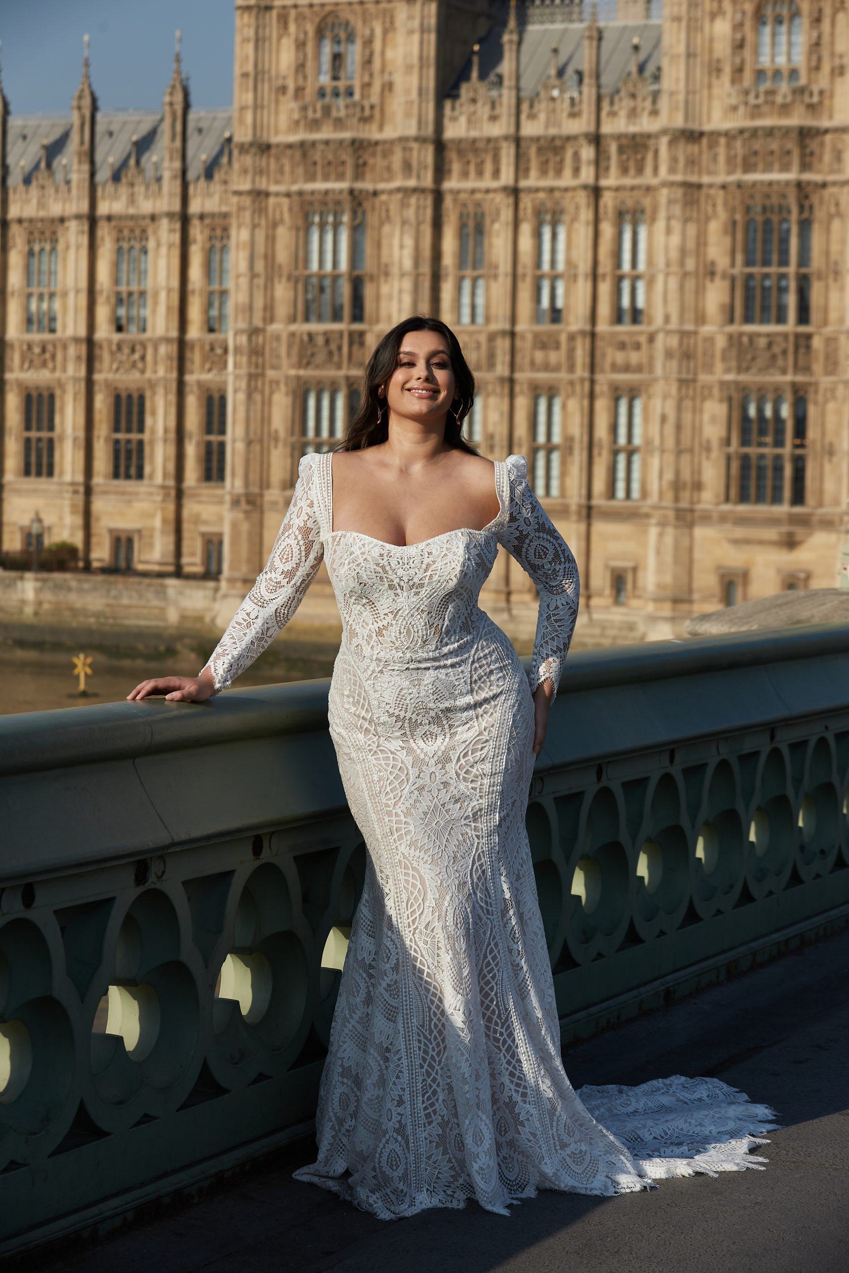 Bride in an Everleigh wedding dress with intricate lace, a sweetheart neckline, and detachable long sleeves, standing on a bridge with a historic building in the background.
