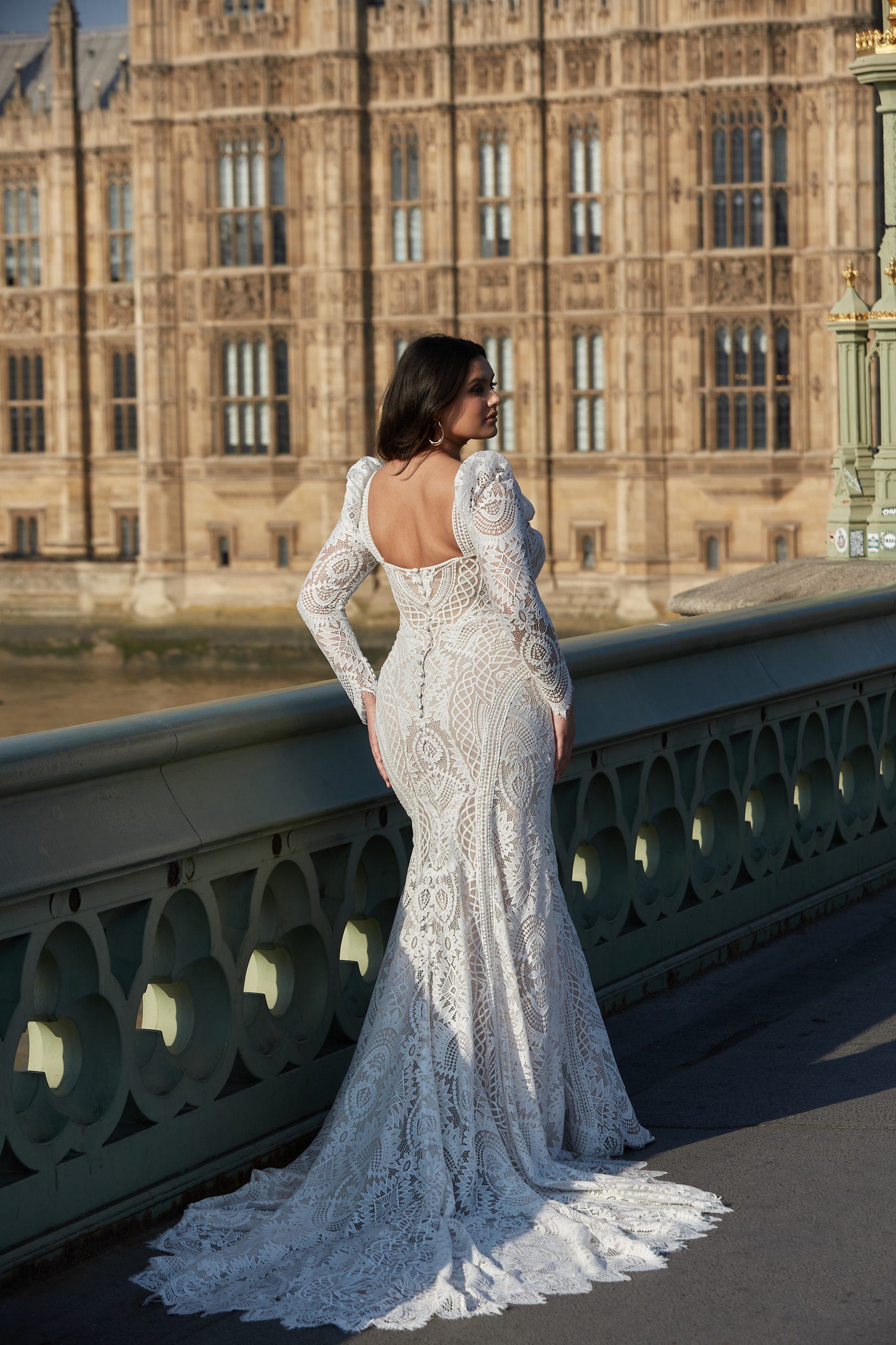 A bride from the Dotty Bridal Collection stands gracefully on Westminster Bridge, overlooking the river with the iconic Houses of Parliament as her backdrop. She is adorned in a stunning, full-length lace gown with long sleeves and a low back that accentuates her figure. The intricate lace patterns flow seamlessly down the dress, ending in a subtle train. Her dark hair cascades down her back, complementing the elegance of the gown. The setting sun casts a warm glow, highlighting the detailed craftsmanship of her dress and the historic architecture behind her.