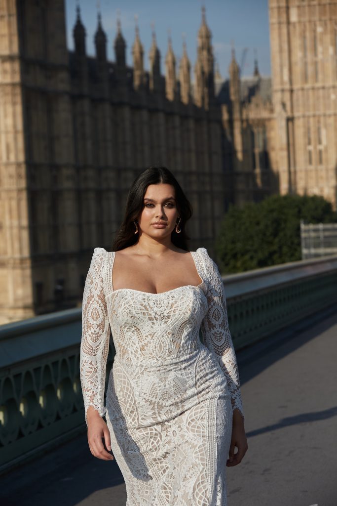 A bride from the Dotty Bridal Collection stands confidently on Westminster Bridge with the iconic Houses of Parliament in the background. She is wearing a form-fitting, long-sleeved gown adorned with intricate lace detailing. The dress features a square neckline that accentuates her décolletage and structured shoulders, adding a touch of modern elegance. Her dark hair flows freely, framing her face, and she wears subtle, classic earrings. The urban, historic setting enhances the timeless beauty of her bridal look, creating a striking and memorable image.