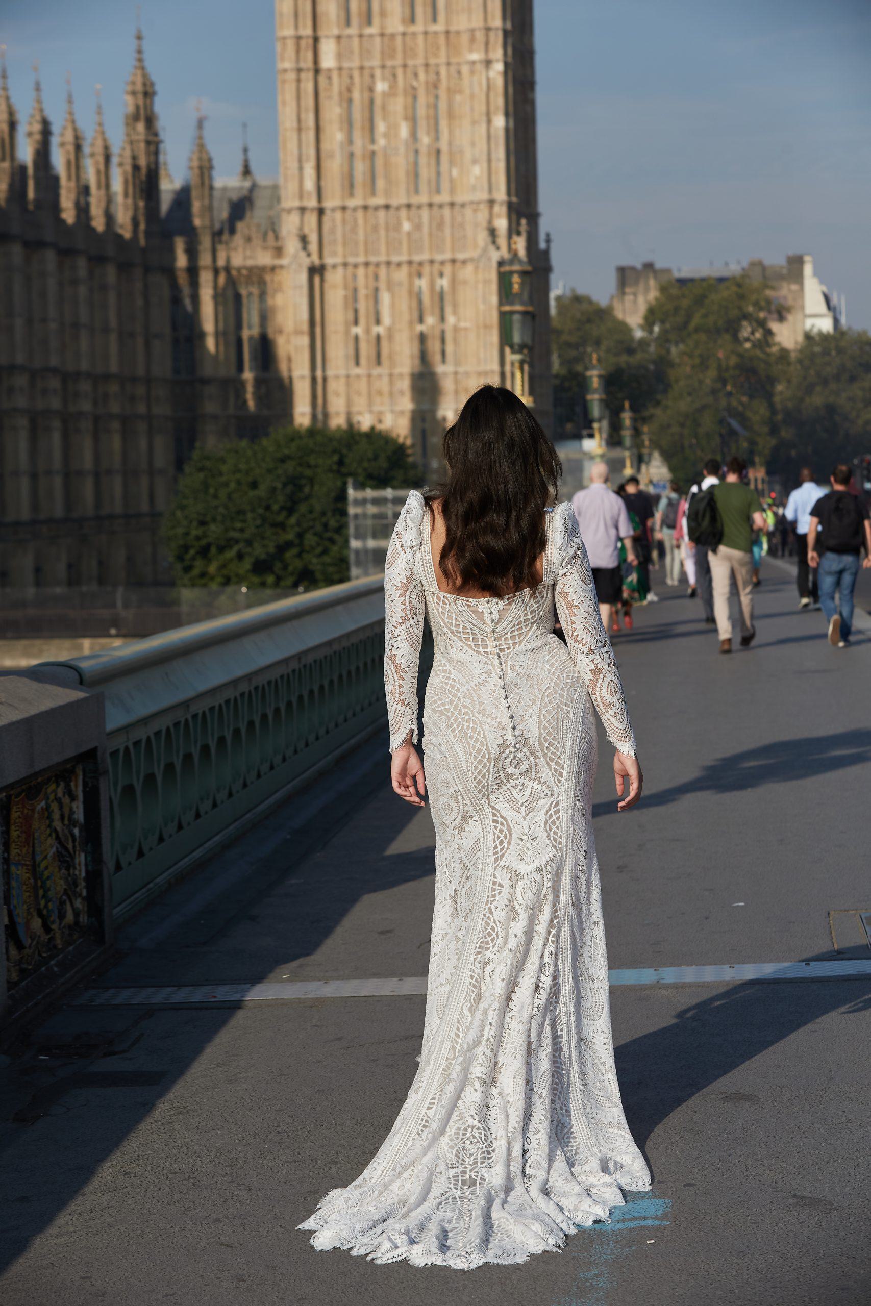 A bride from the Dotty Bridal Collection is seen walking on Westminster Bridge with the iconic Houses of Parliament in the background. She wears a fitted, long-sleeved gown made from intricate lace fabric, accentuating her figure. The dress features a deep square back and a slight train that gracefully trails behind her. Her dark hair falls naturally down her back, and the urban setting creates a striking contrast with the timeless elegance of her wedding dress, capturing a blend of modern and classic bridal beauty.