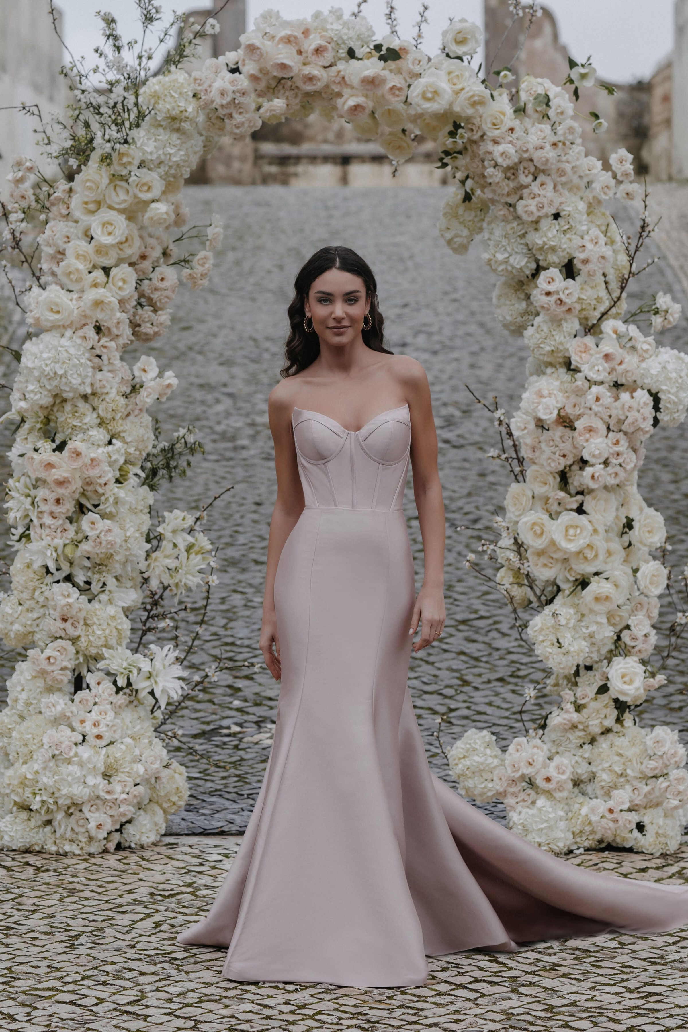 The Sofia by Abella dress is a striking, strapless mermaid gown featuring a structured bodice with prominent seam detailing that accentuates the figure. The gown hugs the waist and hips before flaring out into a sleek, elegant train. The unique blush tone of the dress adds a modern twist, making it a perfect choice for the contemporary bride seeking sophistication and style.