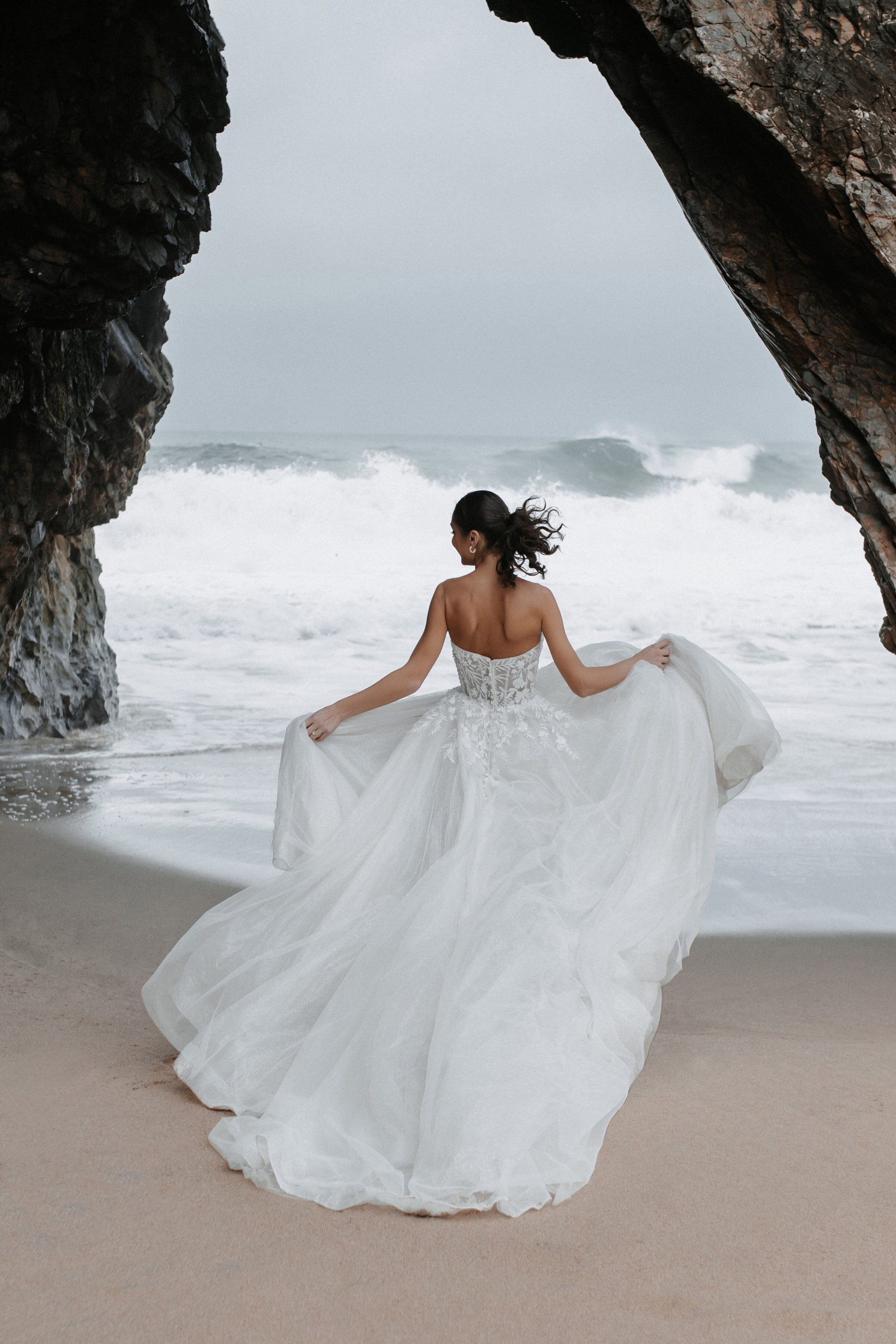 The Benedita by Abella is a captivating strapless gown featuring an intricately beaded bodice that transitions into a voluminous, flowing skirt. The ethereal fabric of the skirt creates a dreamy, sweeping effect, perfect for a romantic beach setting. The back of the dress showcases a delicate lace-up detail, adding a touch of elegance and sophistication.