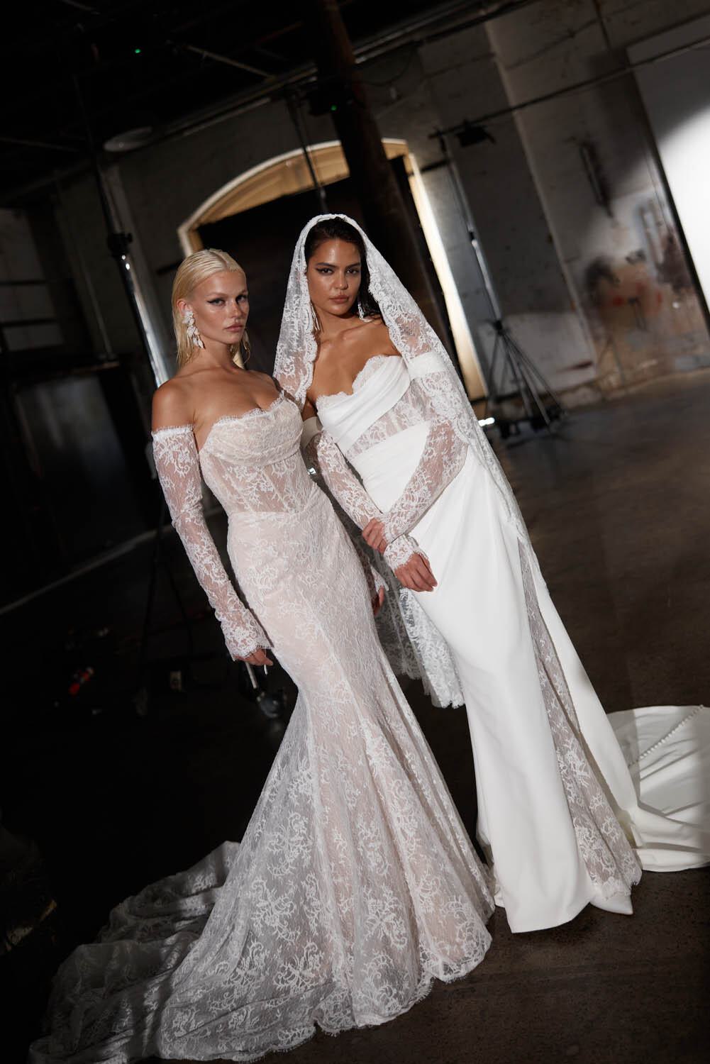 Two models showcasing exquisite contemporary wedding dresses from Evie Young's collection in an industrial setting. The first model wears a form-fitting, off-the-shoulder lace gown with long sleeves and a dramatic train, paired with statement earrings. The second model dons a sleek white gown with an off-the-shoulder neckline, complemented by a lace-trimmed veil and matching long lace sleeves. The dimly lit backdrop with exposed beams and concrete floors enhances the modern and edgy vibe of the collection.