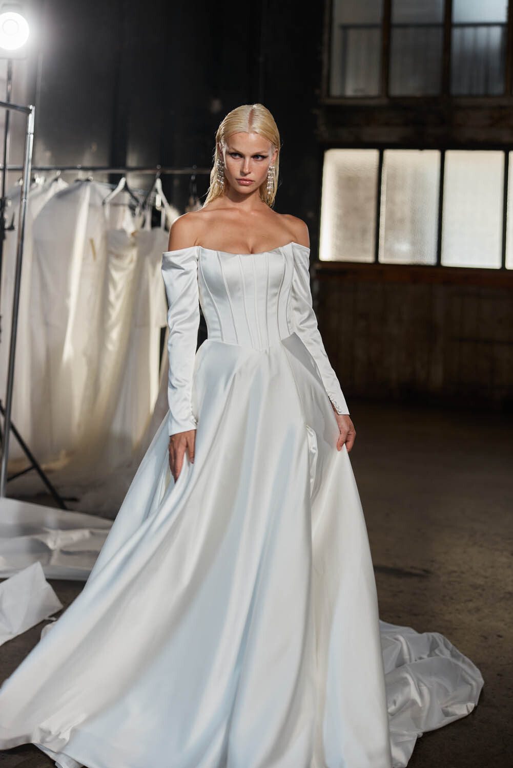 Model showcasing a stunning off-the-shoulder satin bridal gown with a structured corset bodice and long sleeves from Evie Young's collection. The dress features a full, flowing skirt that adds an element of classic elegance. The model stands confidently in an industrial setting with dim lighting and garment racks in the background, highlighting the modern and sophisticated design of the gown.