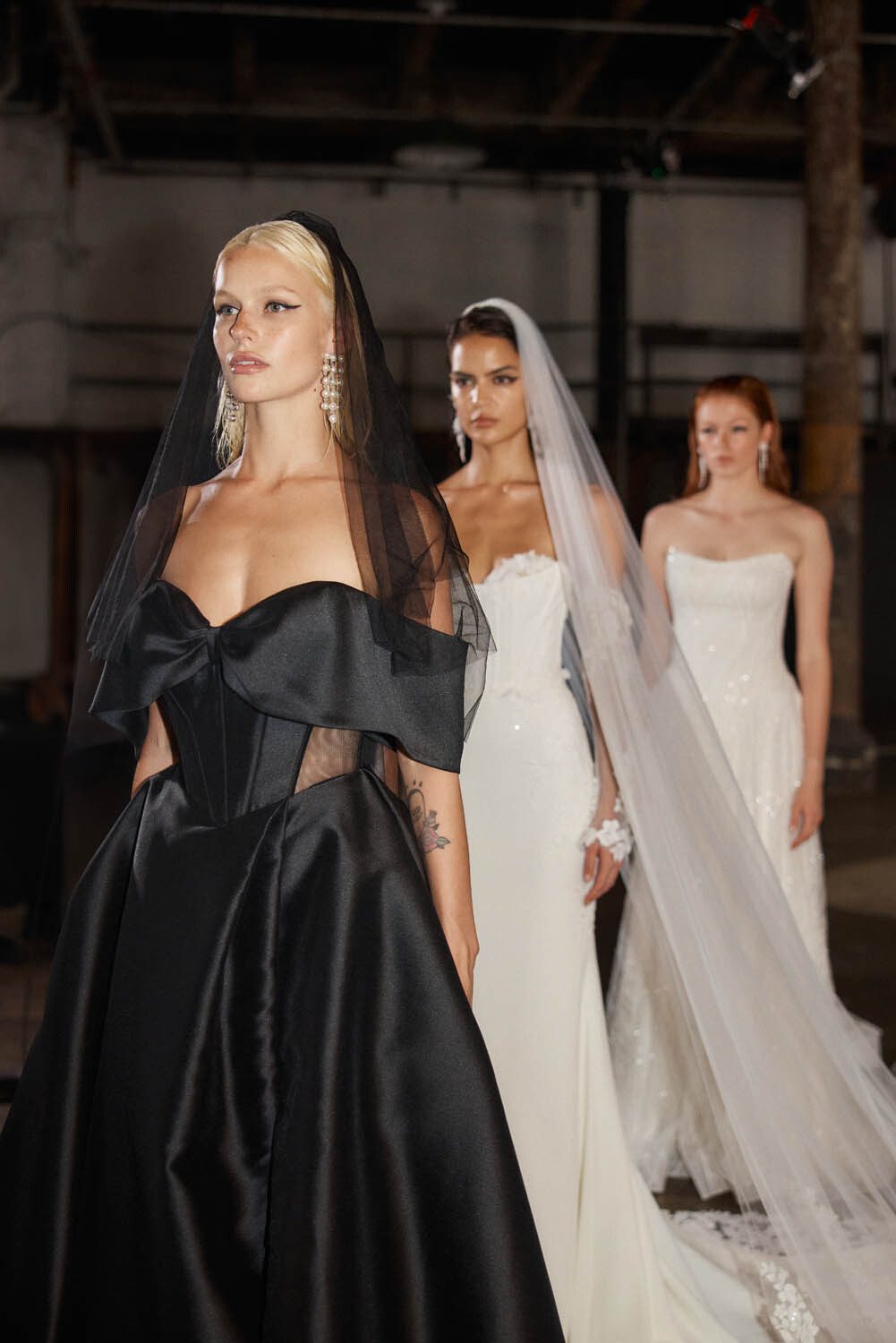 Three models showcasing stunning bridal gowns from Evie Young's collection. The first model wears a dramatic black gown with a structured bodice and sheer elements, complemented by a black veil and statement earrings. The second model dons a sleek, form-fitting white gown with a long train and a delicate veil, exuding timeless elegance. The third model is in a classic white lace gown with a strapless neckline, also with a long veil. The industrial backdrop contrasts beautifully with the sophisticated designs, highlighting the modern and edgy aesthetic of the collection.