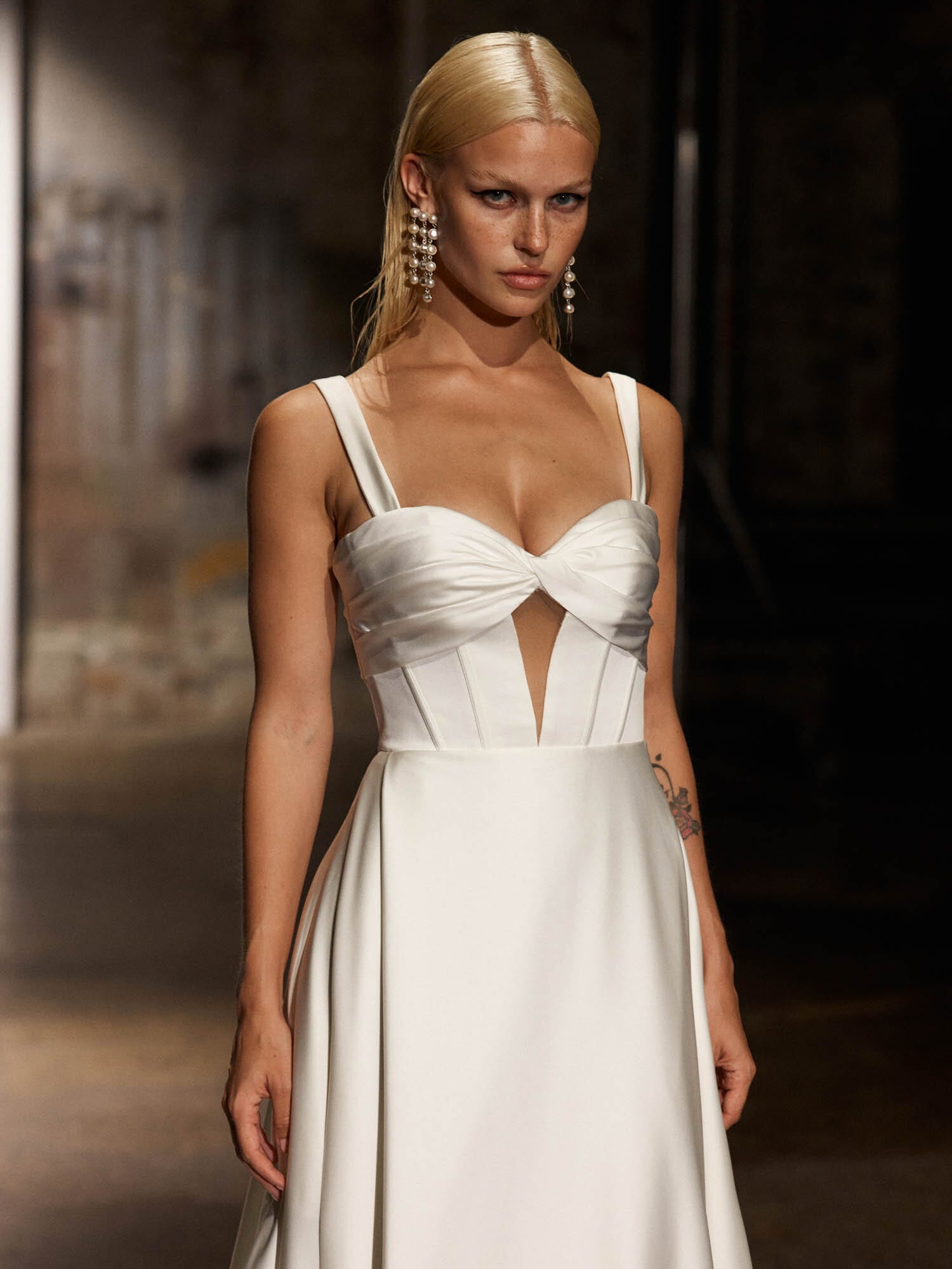 The image showcases a woman in a sleek and elegant white wedding dress. Showing a close up of the front bodice. This gown features a fitted bodice with a keyhole cutout just below the bust, adding a touch of modern style. The dress has wide shoulder straps and a plunging neckline, offering a sophisticated and contemporary look. The skirt flows gracefully to the floor, creating a subtle train that adds to the dress's elegance. The woman is standing in an indoor setting with artistic elements and soft lighting that highlights the smooth, flowing fabric of the dress. The overall impression is one of refined simplicity and modern bridal fashion.
