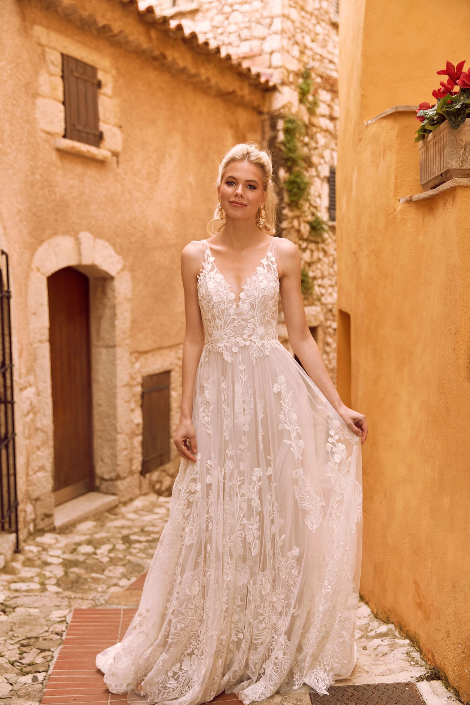 The image shows a woman wearing an elegant and romantic white wedding gown. The dress features a deep V-neckline with delicate lace detailing that extends to the bodice and straps, creating a refined and feminine look. The bodice is fitted, accentuating the woman's figure, and transitions into a flowing, A-line skirt. The skirt is made of light, airy fabric adorned with intricate lace patterns, adding texture and visual interest to the gown. The woman's hair is styled in a simple, elegant updo, complementing the graceful and ethereal design of the dress. The overall effect is one of timeless beauty and romantic elegance, perfect for a fairy-tale wedding.
