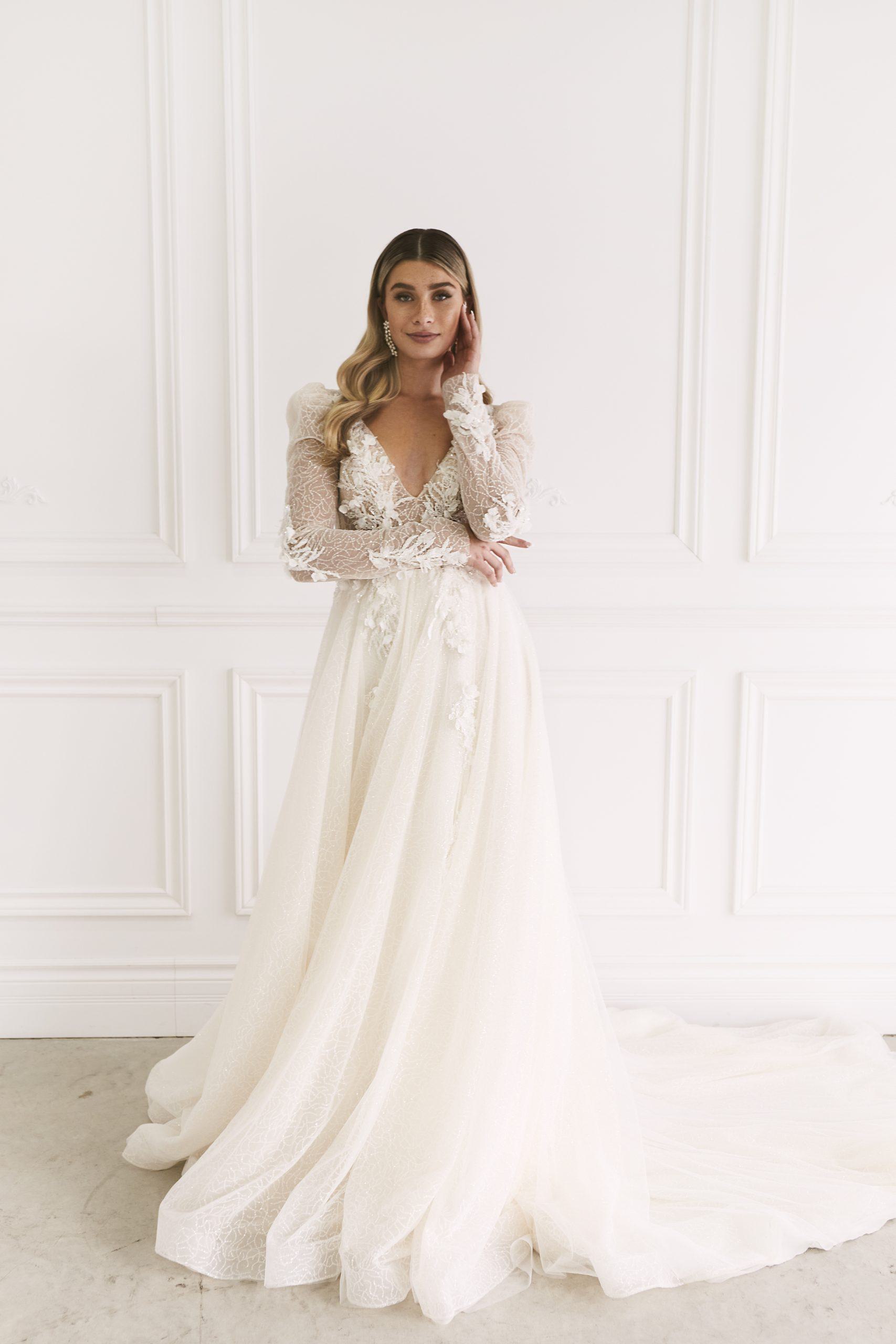 A bride stands gracefully in a luxurious room, wearing a stunningly modern wedding dress that features a deep V-neckline and long, sheer sleeves adorned with intricate lace detailing. The bodice is beautifully fitted, leading into a flowing, full skirt that cascades elegantly to the floor. Her loose waves frame her face, and she gently touches her hair, exuding a timeless and sophisticated elegance.