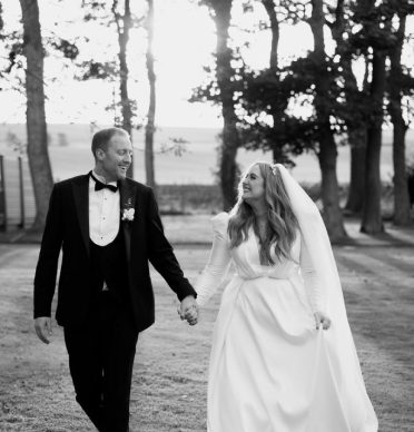 A joyful bride and groom walk hand in hand through a serene, tree-lined outdoor setting, captured in a timeless black-and-white photograph. The bride, dressed in a long-sleeved gown with a flowing veil, looks lovingly at her groom, who is attired in a classic black tuxedo. The soft light filtering through the trees adds a romantic, dreamy quality to the scene, highlighting the couple's happiness as they share this intimate moment together. The natural backdrop of tall trees and open fields provides a peaceful and elegant atmosphere for this special occasion.