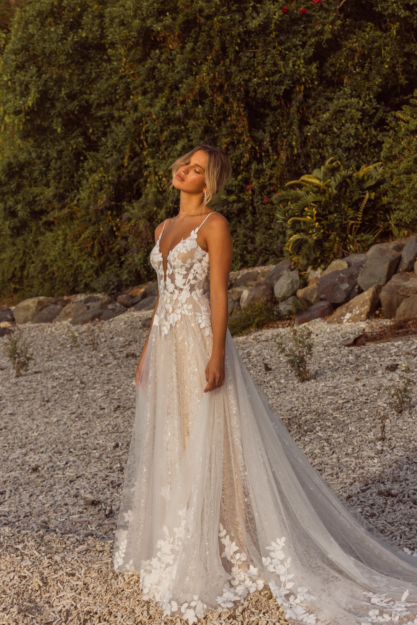 A woman wearing a sleeveless wedding dress with floral lace detailing on the bodice and a sheer overlay skirt. The dress has thin straps and a V-neckline. She is standing and looking slightly upward, with her hair styled in an elegant updo. The dress features a long, flowing train.