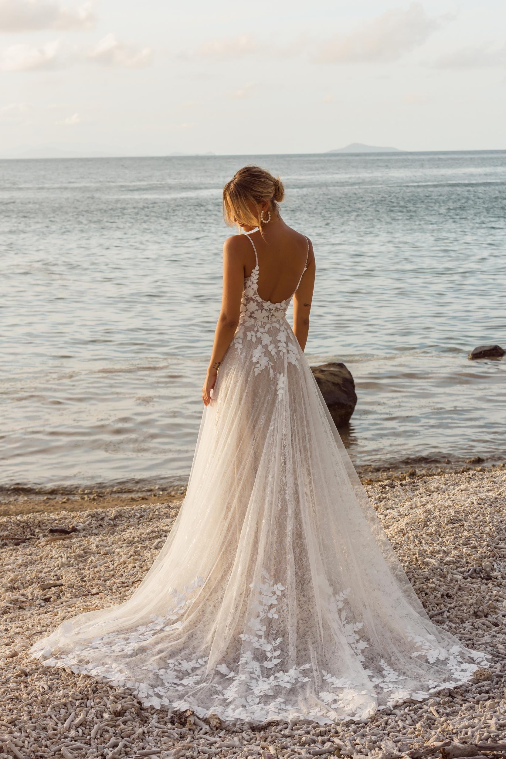 A woman wearing a sleeveless wedding dress with thin straps and a low back. The dress features intricate floral lace detailing on the bodice and a long, flowing skirt with a sheer overlay and lace accents extending into a train. Her hair is styled in an elegant updo, and she is standing, facing away, showcasing the back of the dress.