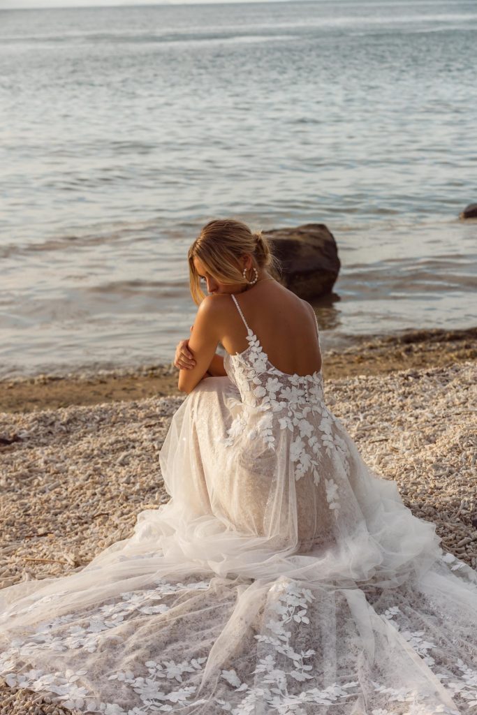 A woman wearing a sleeveless wedding dress with a low back and lace detailing, sitting with her back to the camera. Her hair is styled in an updo.