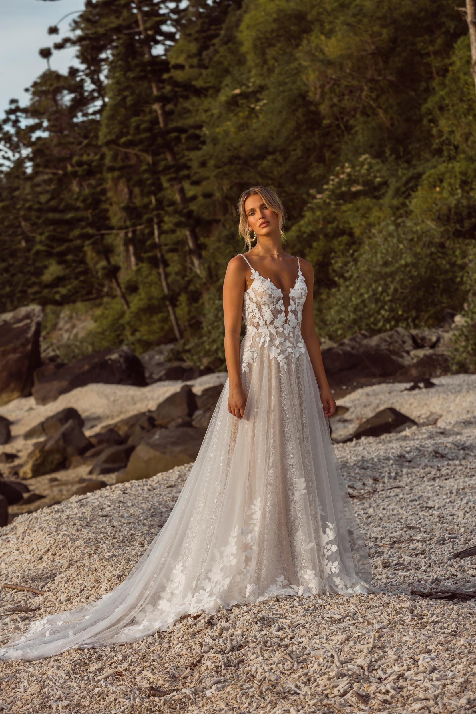 A woman wearing a sleeveless wedding dress with a fitted bodice adorned with intricate lace detailing and a flowing train.