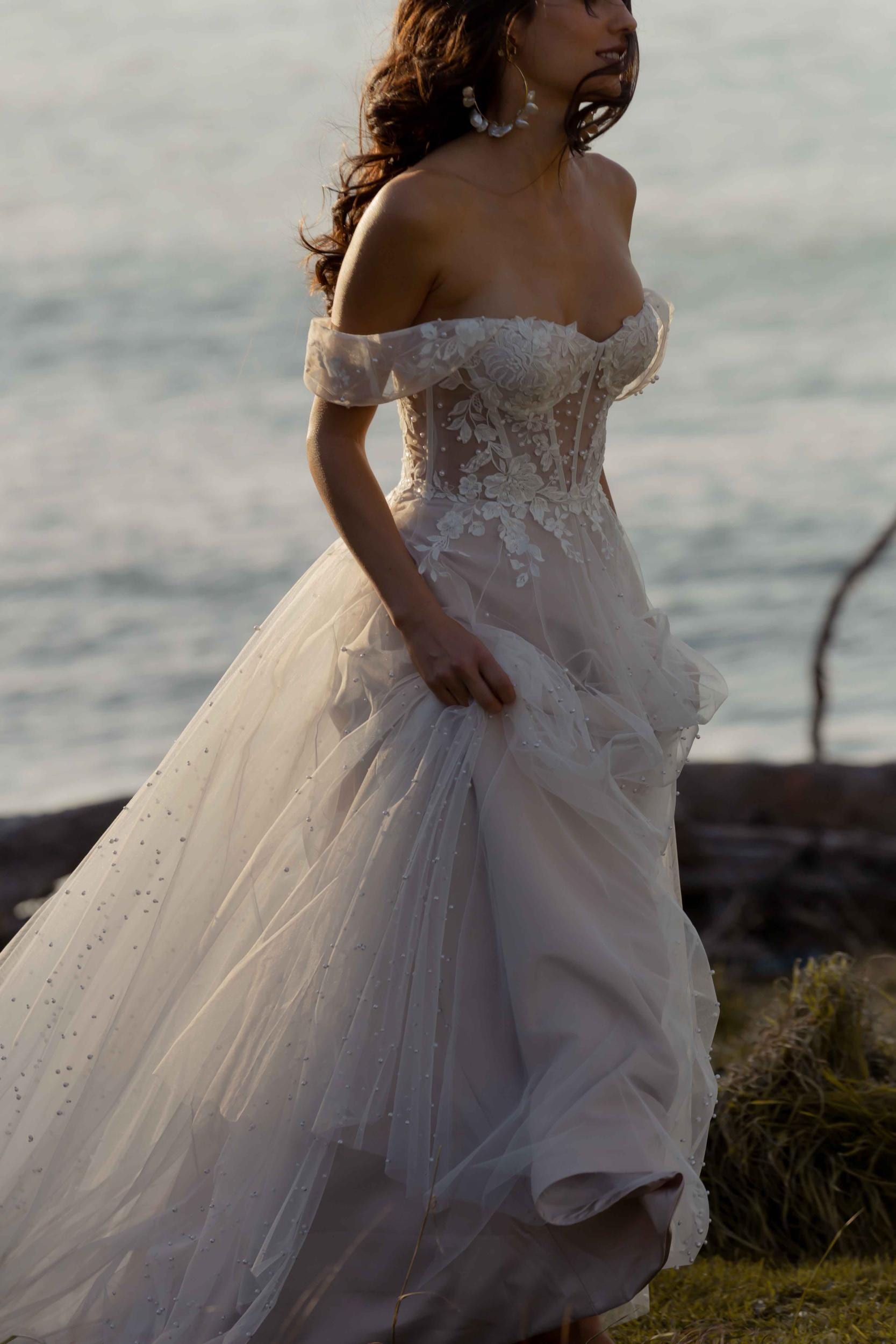 The image shows a woman wearing a stunning wedding gown with an off-the-shoulder design. The bodice is fitted and features intricate lace and beadwork detailing, adding texture and elegance. The gown has a voluminous skirt made of layers of tulle, creating a full and flowing silhouette. The fabric appears light and airy, contributing to the gown's romantic and ethereal quality. The woman's hair is styled in loose waves, cascading over her shoulders, complementing the overall romantic and elegant design of the dress. The overall effect is one of timeless beauty and refined elegance, perfect for a stylish and sophisticated wedding.