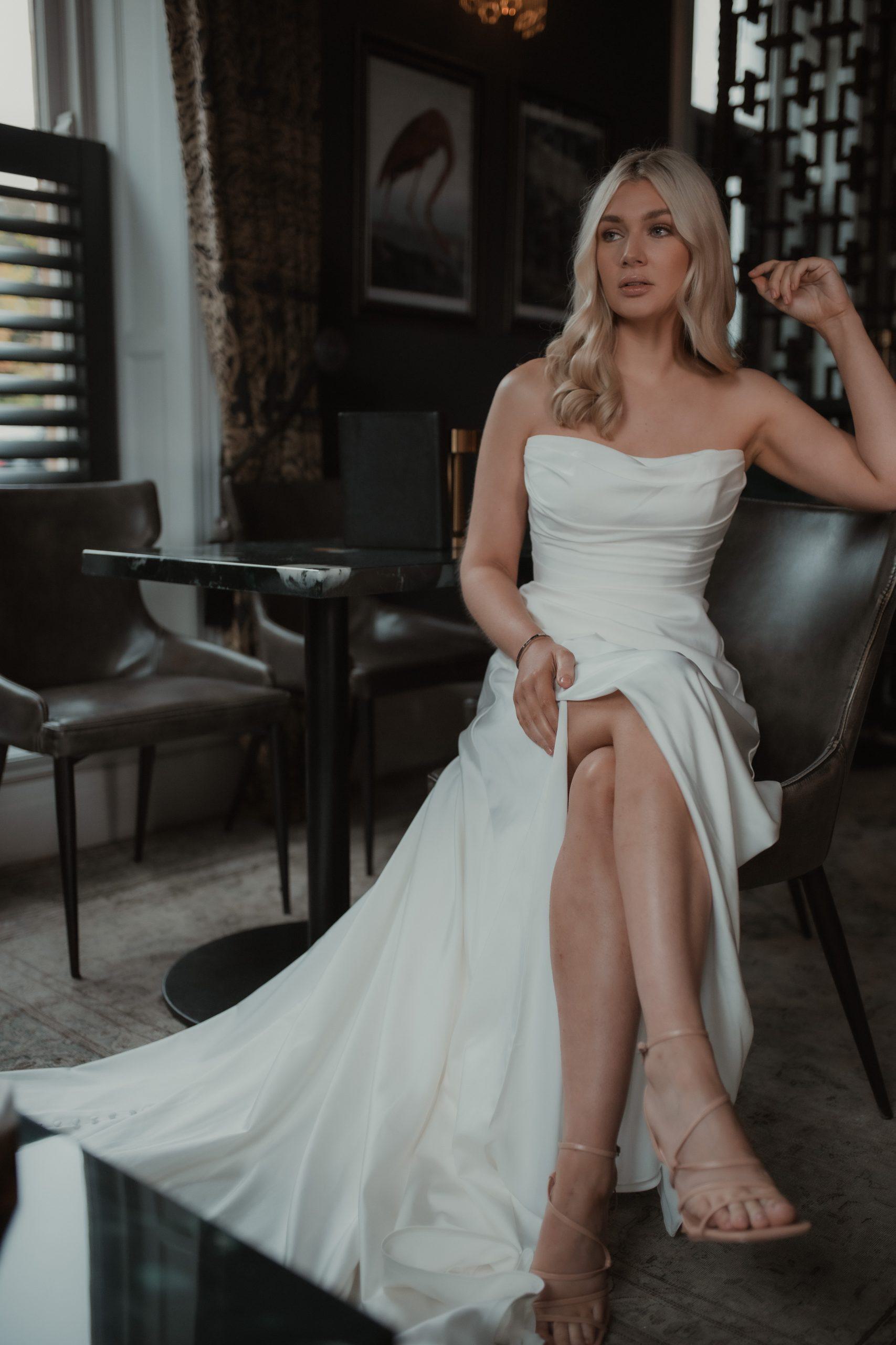 A bride sits elegantly in a sophisticated indoor setting, wearing a strapless wedding gown with a ruched bodice and a flowing skirt. The dress features a thigh-high slit, showcasing her strappy heels. Her blonde hair is styled in loose waves, and she has a serene and thoughtful expression. The background includes stylish furnishings, framed artwork, and soft lighting, creating an intimate and chic ambiance.