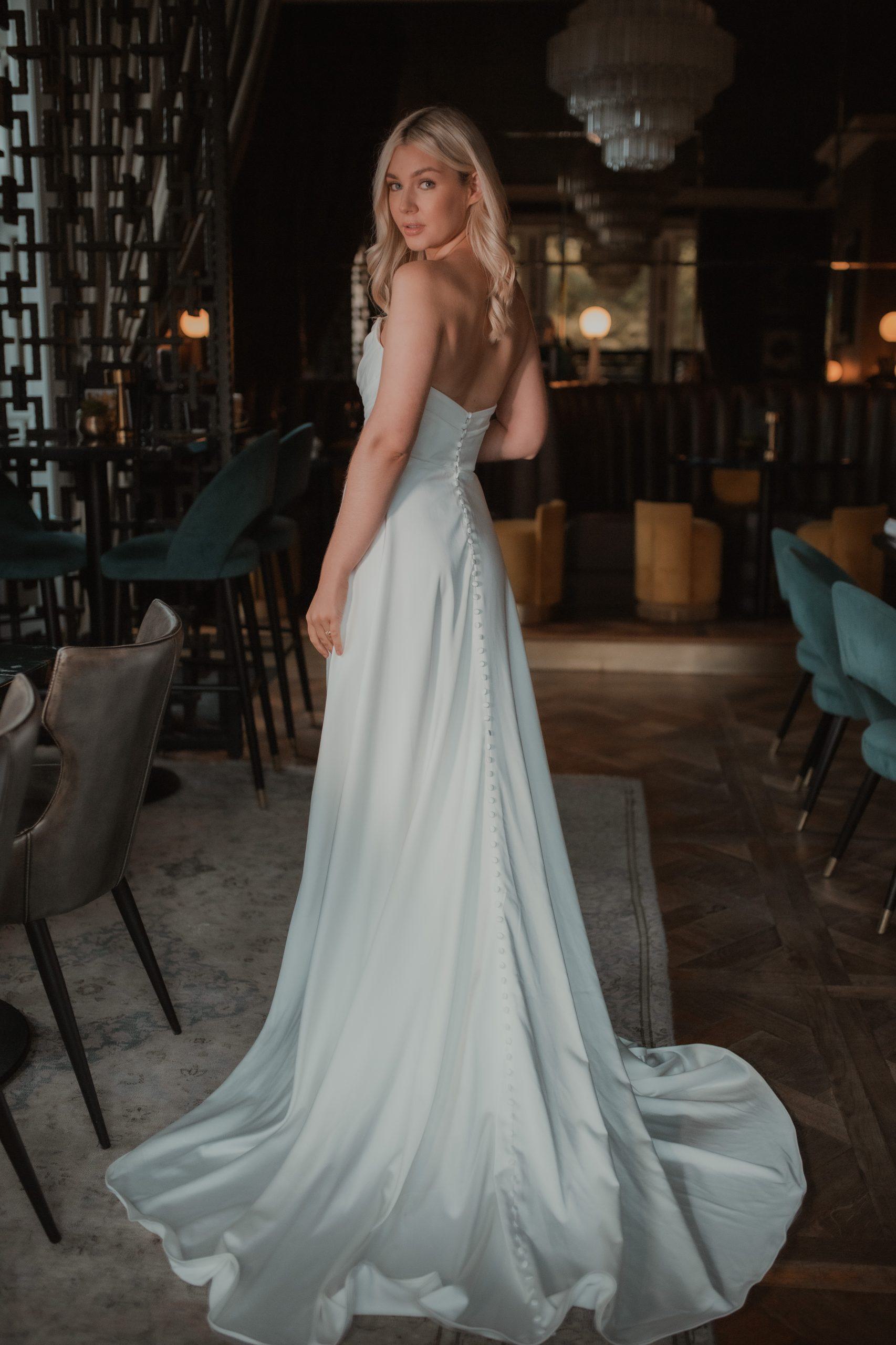 A bride stands confidently in a luxurious strapless wedding gown with a fitted bodice adorned with intricate lace detailing. The dress features a full skirt that cascades elegantly to the floor, creating a timeless and romantic silhouette. The bride's hair is styled in loose waves, complementing her natural beauty as she gazes thoughtfully to the side. The background is dark and intimate, with subtle lighting highlighting the bride and the exquisite details of her gown.