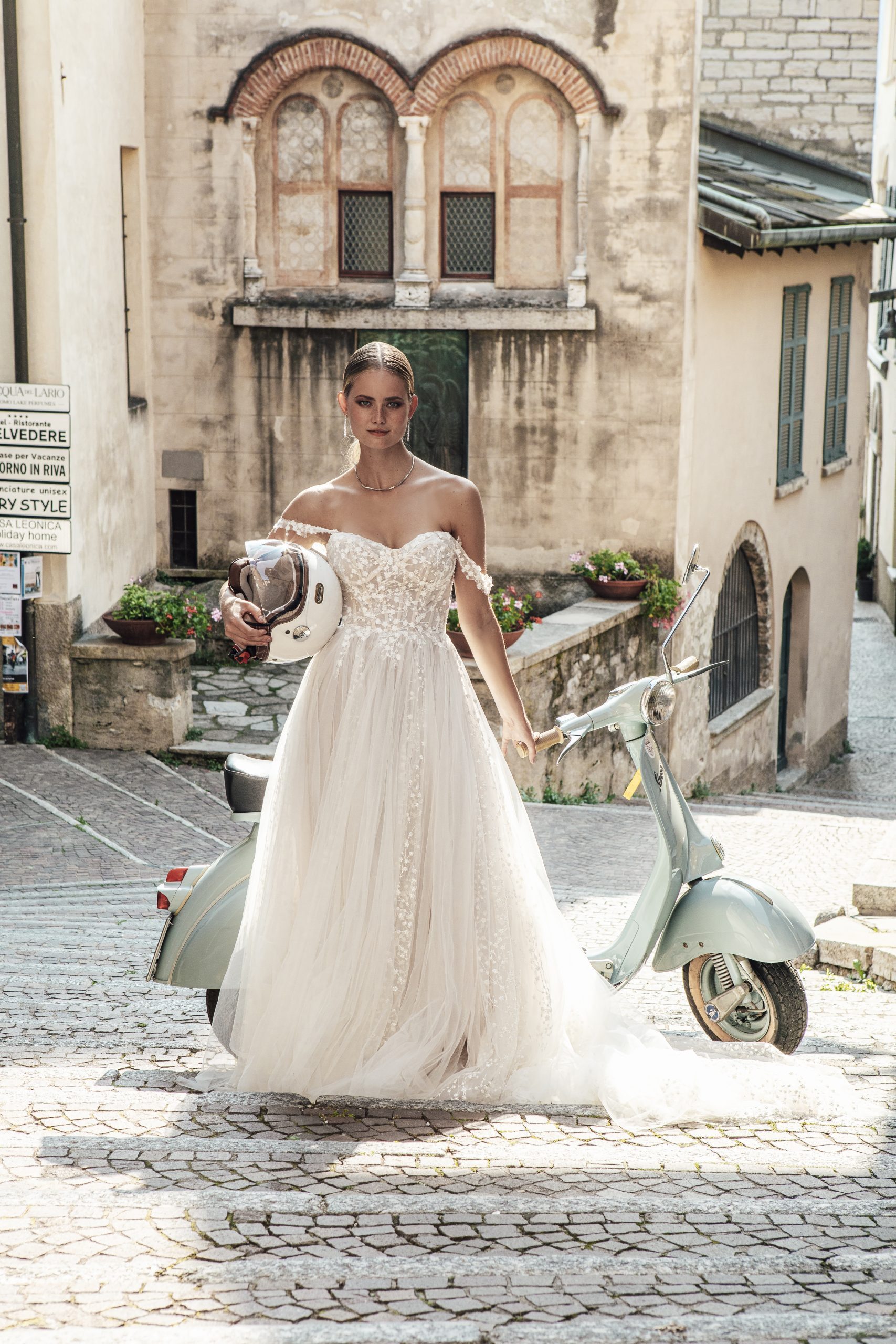 A bride stands in the Hopkins gown, a flowing off-the-shoulder dress adorned with intricate lace and beadwork that adds a touch of romantic elegance. She holds a vintage helmet in one hand, perfectly complementing the pastel Vespa scooter parked beside her. The cobblestone street and historic architecture in the background evoke a timeless, European charm, while the gown’s ethereal train softly trails behind her, blending tradition with a modern, adventurous spirit.