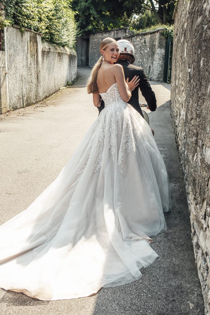 A bride is seated on the back of a scooter, holding onto the groom, who is dressed in a suit and helmet, ready to ride. She is wearing a stunning Hilton gown, featuring a strapless bodice with intricate lace detailing that flows into a voluminous skirt with a long, trailing train. The bride looks back over her shoulder with a bright smile, her hair styled in a sleek ponytail. The scene is set on a narrow, sunlit street with high stone walls, capturing a joyful and spontaneous moment as the couple embarks on their next adventure together.