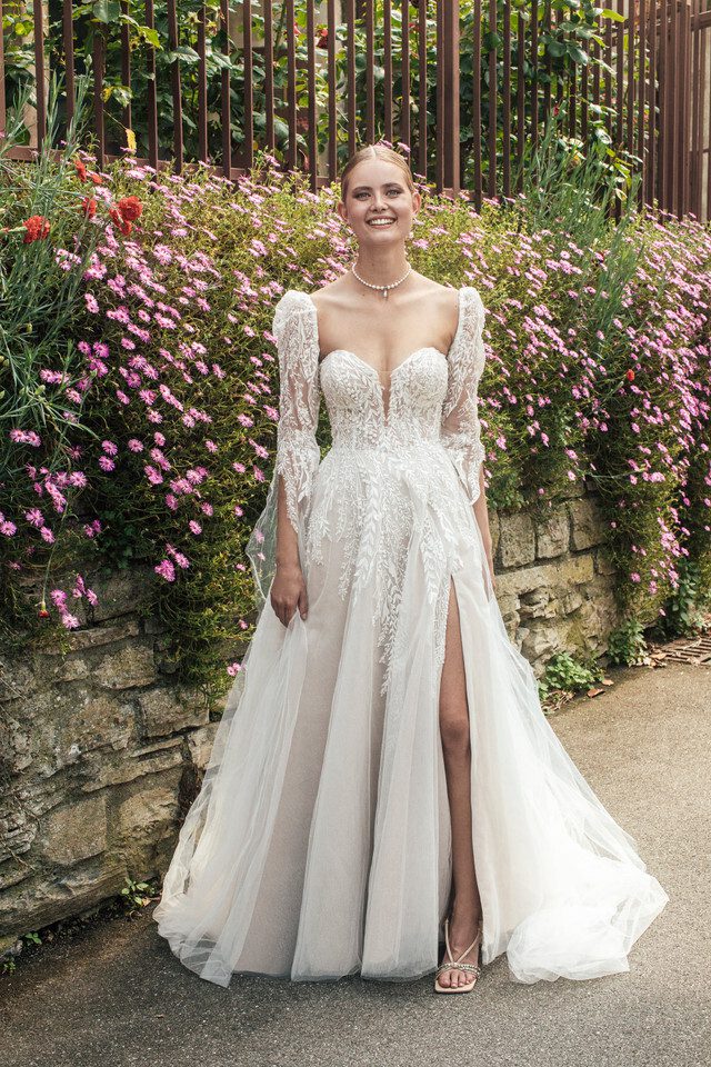 A bride stands confidently in a garden, wearing an exquisite gown with a plunging sweetheart neckline and intricately detailed lace bodice. The dress features long, sheer sleeves adorned with delicate lace appliqués and a high slit that adds a modern touch to the flowing tulle skirt. The bride’s radiant smile is framed by a simple yet elegant updo, and she accessorizes with a pearl necklace. The backdrop of blooming flowers and greenery enhances the romantic and ethereal atmosphere of the scene, perfectly complementing the beauty and elegance of the bridal ensemble.