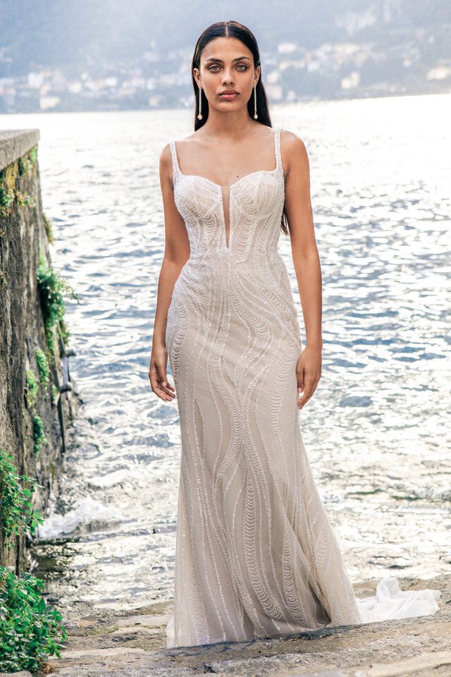 A bride stands confidently in the Huntleigh gown, a stunning piece that features a deep plunging neckline with delicate straps. The gown is adorned with intricate beadwork that cascades down the length of the dress in a mesmerizing wave pattern, echoing the serene waters behind her. The fitted silhouette enhances her figure, while the natural setting of stone steps and rippling water creates a romantic, almost ethereal atmosphere. Her poised expression and the gown's exquisite detailing capture the essence of timeless elegance.