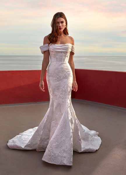 The image features the Gloria wedding gown, an elegant off-the-shoulder mermaid dress with a fitted, corset-style bodice and a full, flared skirt that extends into a subtle train. The gown is adorned with delicate floral patterns that add texture and depth. The bride stands against a minimalist backdrop with a soft, pastel-hued sky and a red curved wall, creating a striking contrast that emphasizes the gown's sophisticated design. Her hair falls in loose waves, complementing the romantic and timeless feel of the dress.