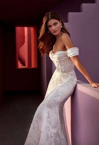 The image shows a bride wearing the Gloria wedding gown, a stunning off-the-shoulder mermaid dress that features a structured bodice with delicate floral embroidery. The gown hugs her figure and flows elegantly down to the floor. She is seated on a pastel-colored ledge, with modern architectural elements in bold reds and purples surrounding her, creating a contemporary and artistic atmosphere. The bride's hair is styled in loose waves, adding a soft, romantic touch to the overall sophisticated look.