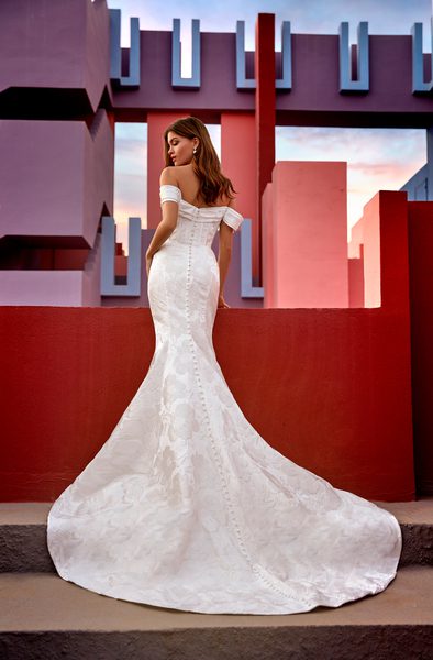 The image features a bride standing with her back to the camera, wearing an off-the-shoulder mermaid-style wedding gown named Gloria. The gown is fitted through the bodice and hips, then flares out into a dramatic train, beautifully showcasing the intricate floral lace pattern that flows down the length of the dress. The backdrop is a striking architectural setting in bold red and blue tones, adding a modern, artistic flair to the image. The bride's hair is styled in loose waves, cascading down her back, complementing the elegance and sophistication of the gown.
