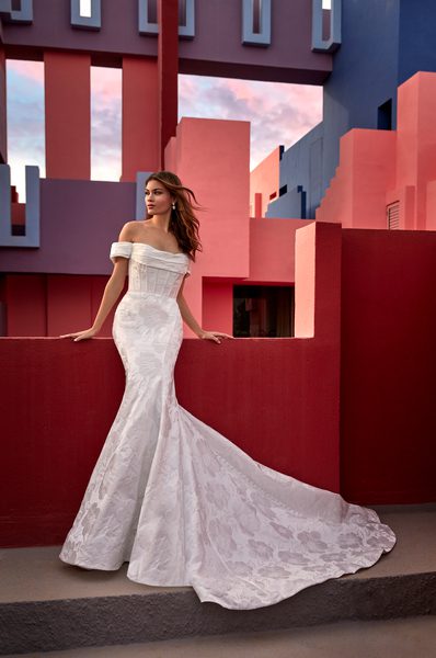 The image features a bride standing against a vibrant backdrop of geometric architecture in bold shades of red and blue. She wears a stunning off-the-shoulder wedding gown that hugs her figure before flaring out into a dramatic train. The dress is adorned with a subtle floral lace pattern, adding texture and elegance to the overall look. Her hair is styled in loose waves that cascade over her shoulders, complementing the gown's romantic yet modern style. The contrast between the classic bridal look and the contemporary architectural setting creates a striking visual appeal.