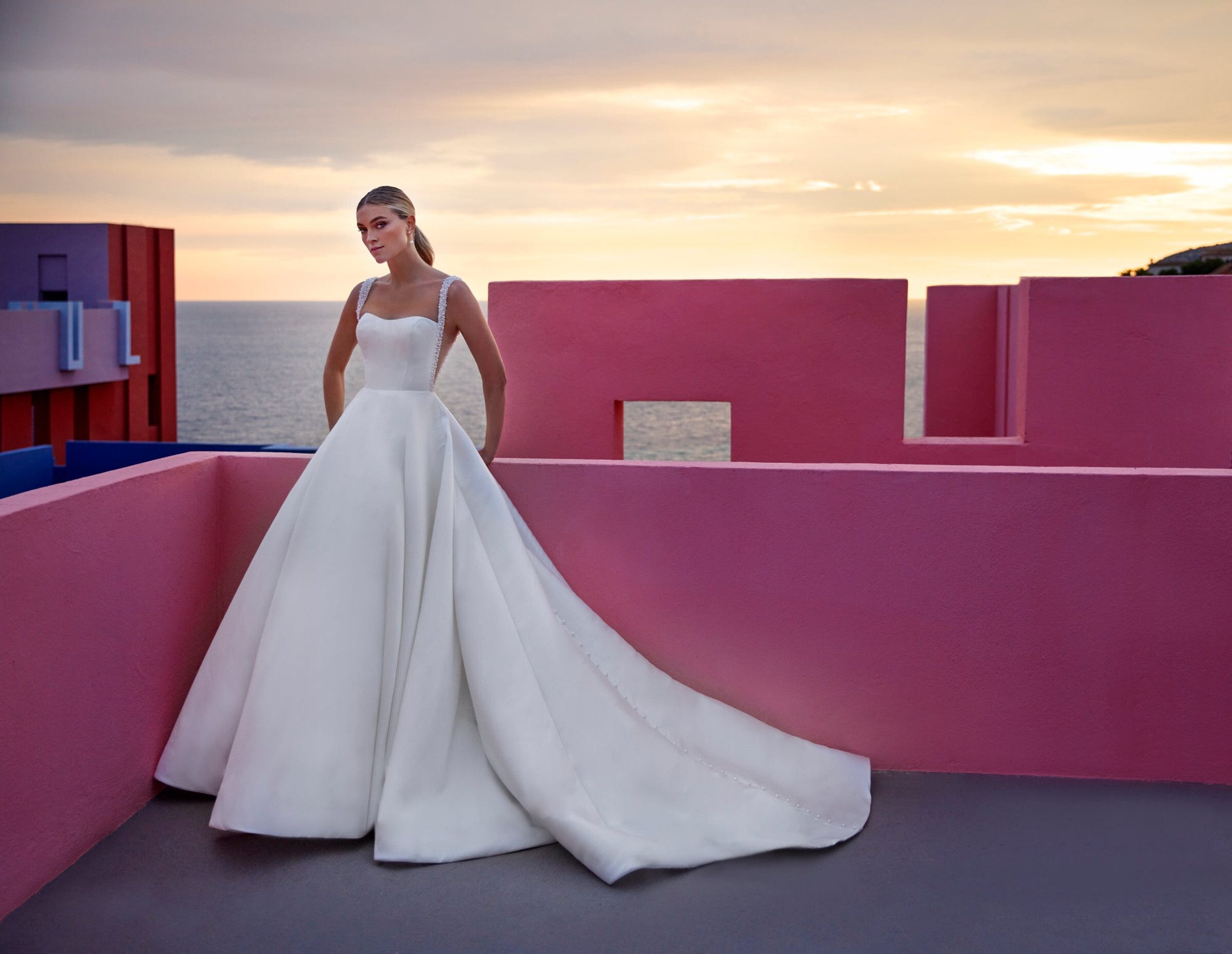 A bride poses against a vibrant pink wall at sunset, wearing a stunning Madison James wedding gown. The dress features a fitted bodice with a sweetheart neckline and intricately beaded straps. The full, voluminous skirt flows into a dramatic train, creating a striking silhouette. Her hair is elegantly styled, complementing the modern yet timeless elegance of the gown. The backdrop of the ocean and the warm hues of the sunset add a romantic touch to the scene.