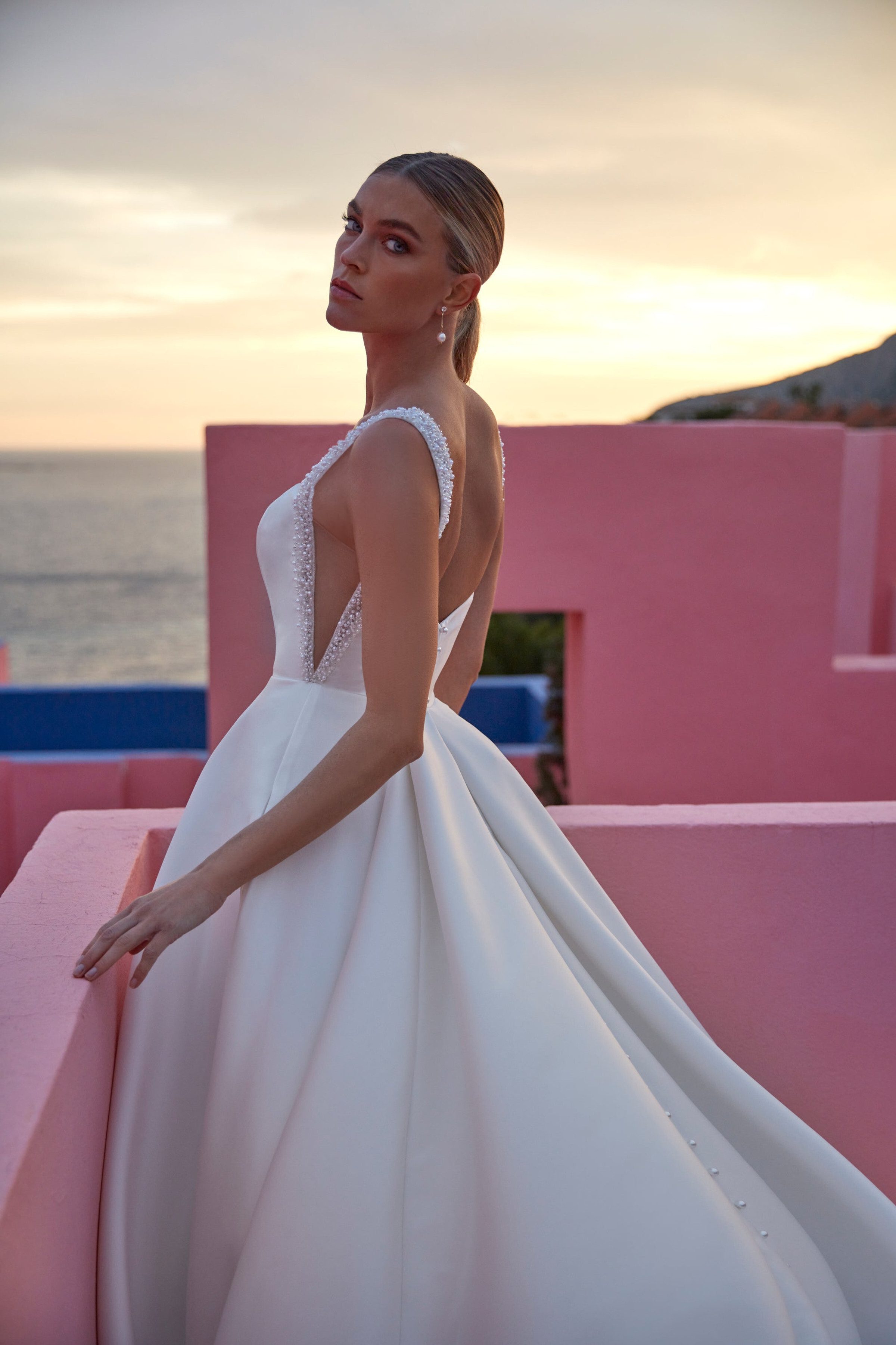 A bride stands against a pastel pink wall at sunset, wearing an exquisite Madison James wedding dress. The gown features a deep V-back adorned with delicate beading along the edges. The bodice has sheer side panels that add a modern twist to the classic silhouette. The full satin skirt flows gracefully, highlighted by a subtle row of buttons cascading down the back. Her hair is elegantly pulled back, complementing the sophisticated and timeless appeal of the dress.