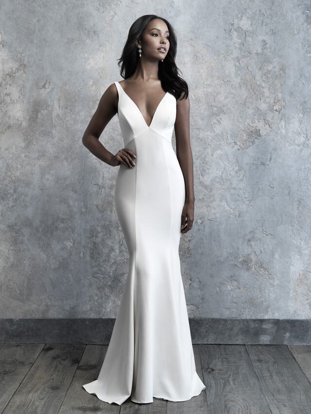 The image showcases a bride wearing a sleek, minimalist wedding gown that hugs her figure gracefully. The dress features a deep V-neckline and wide straps that complement the fitted bodice, seamlessly flowing into a mermaid silhouette that gently flares out at the bottom. The gown's fabric is smooth and slightly reflective, highlighting the bride's elegant form. She stands confidently against a textured gray background, with one hand resting on her hip. Her dark hair cascades in soft waves, and she wears delicate drop earrings, completing the look with poise and sophistication. The overall impression is one of modern elegance and timeless beauty.