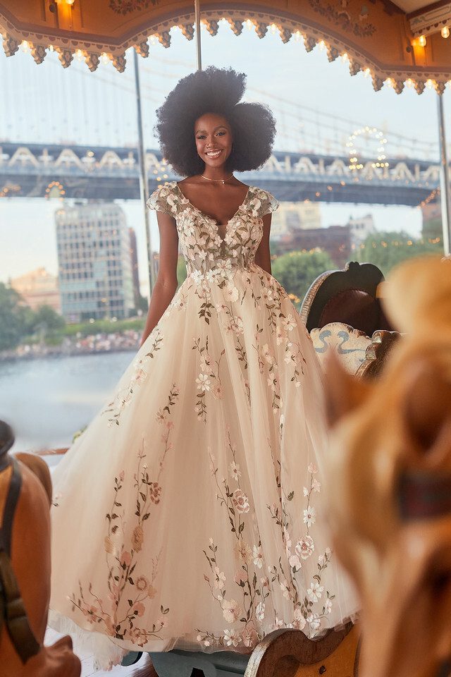 A bride stands gracefully on a vintage carousel, wearing a dreamy blush-colored ball gown embellished with delicate floral appliqués. The gown features a deep V-neckline and cap sleeves, exuding a romantic and ethereal charm. Her voluminous afro frames her smiling face, adding to the whimsical and joyful atmosphere. In the background, the iconic structure of a bridge and the city skyline are visible, blending the magical, fairytale-like setting with the urban environment. The soft lighting and intricate details of the carousel enhance the enchanting feel of the scene.
