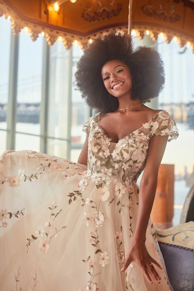 The image features a joyful bride standing in an elegant setting, wearing a breathtaking floral wedding gown. The dress is a soft, blush-colored ball gown adorned with delicate floral appliqués that cover the bodice and scatter down the full tulle skirt. The gown has a deep V-neckline and cap sleeves, with the floral motifs adding a whimsical touch. The bride's natural hair is styled in a voluminous afro, and she beams with happiness, holding the edge of her skirt playfully. The background includes a carousel with glowing lights, enhancing the magical, fairytale-like atmosphere of the scene.