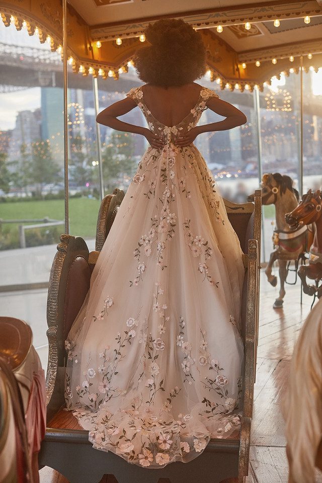 The image shows a bride standing on a carousel ride, captured from behind. She is wearing a stunning, blush-colored ball gown wedding dress adorned with intricate floral appliqués that cascade down the length of the gown. The dress features a deep V-back and cap sleeves, creating an elegant and timeless look. Her voluminous natural afro adds to the overall ethereal vibe of the scene. The carousel's warm lights and vintage details, along with the view of an urban park and city skyline visible through the glass, contribute to the enchanting atmosphere, blending the whimsical with the modern.