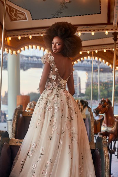The image shows a bride standing on a vintage carousel, looking back over her shoulder with a radiant smile. She wears a romantic, ethereal wedding gown with a deep V-back and delicate, sheer cap sleeves. The gown is adorned with intricate floral appliqués that cascade down the bodice and flow into the full tulle skirt. The soft colors of the floral designs give the dress a whimsical, fairytale-like charm. Her natural afro is styled voluminously, adding to the overall enchanting and joyful atmosphere of the scene. The background features a blurred cityscape with twinkling lights, creating a dreamy contrast to the carousel's ornate details and warm lighting.