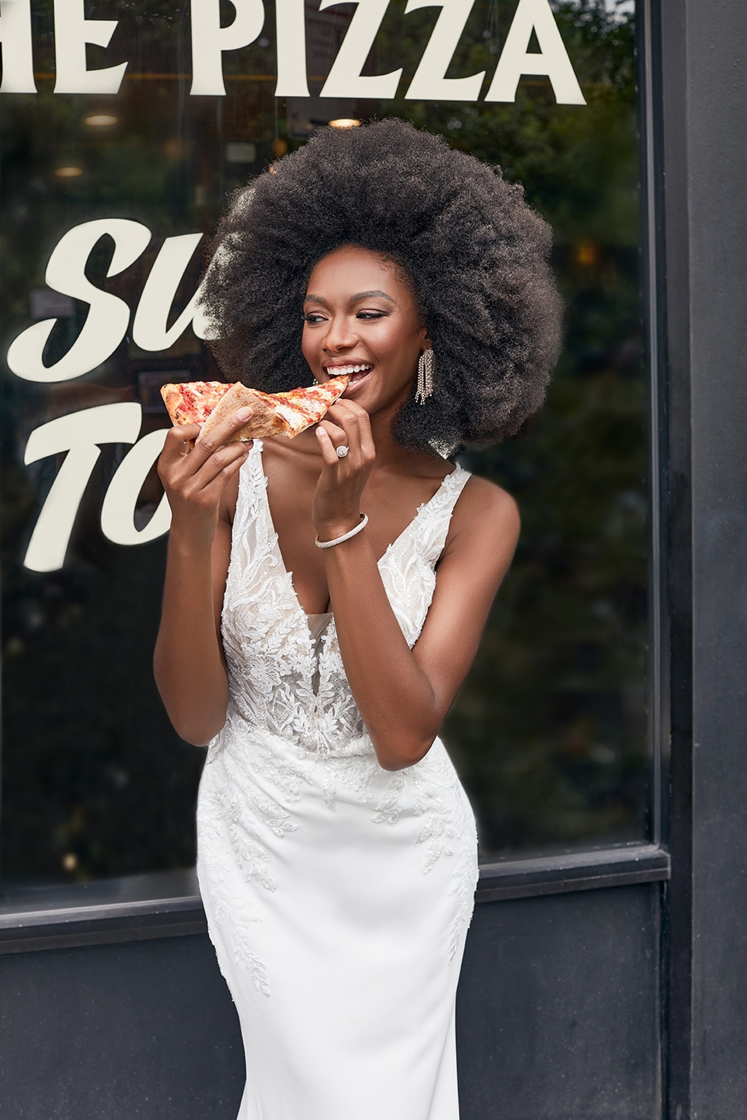 A bride wearing the "Joana" gown by Madison James stands in front of a pizza shop window, joyfully taking a bite of a slice of pizza. The dress features intricate lace detailing on the bodice with a deep V-neckline and a fitted silhouette that highlights her figure. Her voluminous natural curls and radiant smile add a playful and relaxed vibe to the elegant bridal look. The combination of bridal elegance and the casual pizza moment creates a unique and charming scene.
