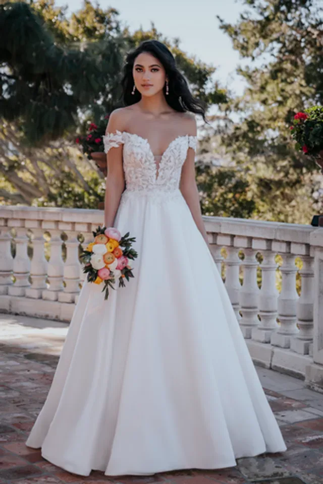 An elegant wedding dress featuring a deep V-neckline adorned with delicate floral lace appliqués. The off-the-shoulder sleeves are crafted from the same lace, adding a romantic touch to the bodice. The lace detailing extends slightly below the waist, where the dress transitions into a full A-line skirt made from smooth, luxurious fabric. This combination creates a timeless, ethereal look perfect for a fairytale wedding.