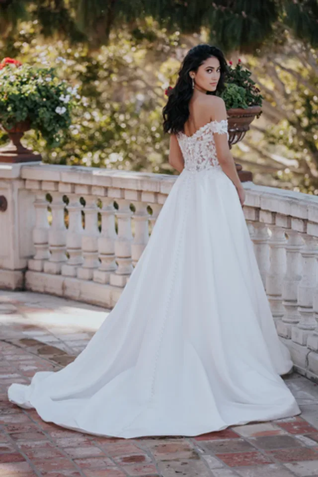 A bride is captured from behind, standing on a sunlit terrace, wearing a stunning wedding gown, style R3660 03. The dress features an off-the-shoulder lace bodice with intricate floral detailing that extends into a fitted waist. The back of the gown showcases a delicate row of buttons that trail down into the full, flowing train, creating a seamless and elegant look. Her dark hair falls in soft waves over her shoulders as she glances back, adding to the romantic and timeless feel of the scene. The lush greenery and ornate stone balustrade provide a perfect backdrop, highlighting the elegance of the gown.