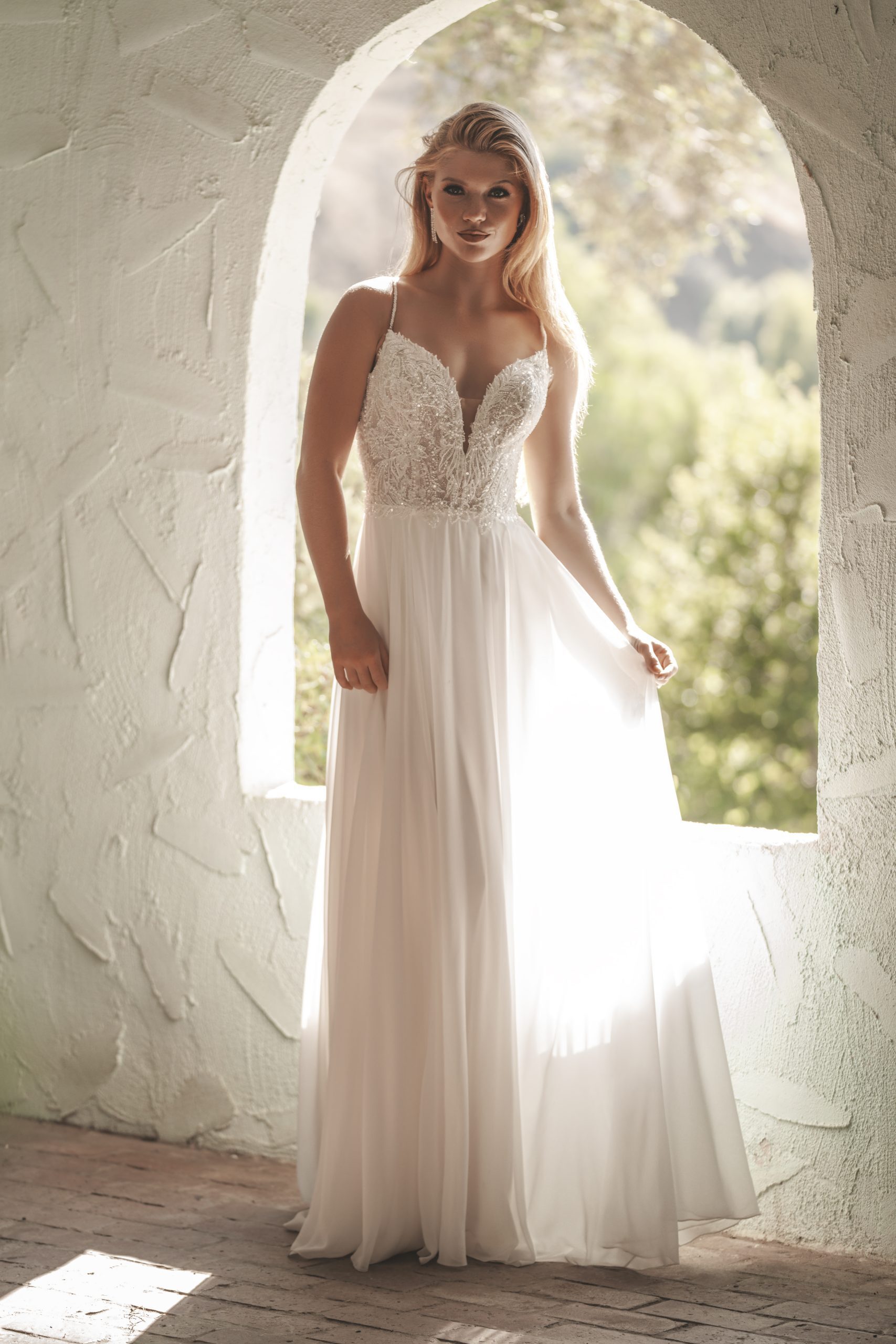 A delicate wedding dress featuring a plunging V-neckline and thin spaghetti straps, adorned with intricate floral lace appliqués on the bodice. The dress flows into a soft, airy chiffon skirt, creating a graceful and romantic A-line silhouette.