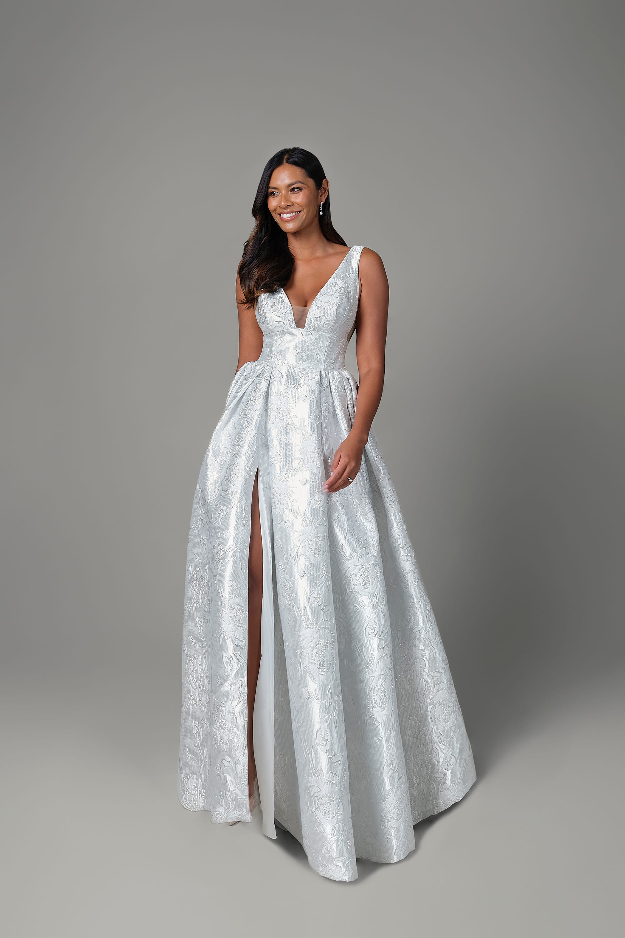 A woman is standing in a minimalist studio setting, wearing a stunning white ball gown with a deep V-neckline and sleeveless design. The gown features a fitted bodice and a full skirt adorned with a subtle floral pattern, creating an elegant and luxurious look. The skirt has a high slit, revealing one leg and adding a modern touch to the classic silhouette. The dress also includes pockets, blending style with practicality. The woman's hair is styled in loose waves, and she is smiling, exuding confidence and grace. The background is plain and neutral, keeping the focus on the elegance and details of the gown.