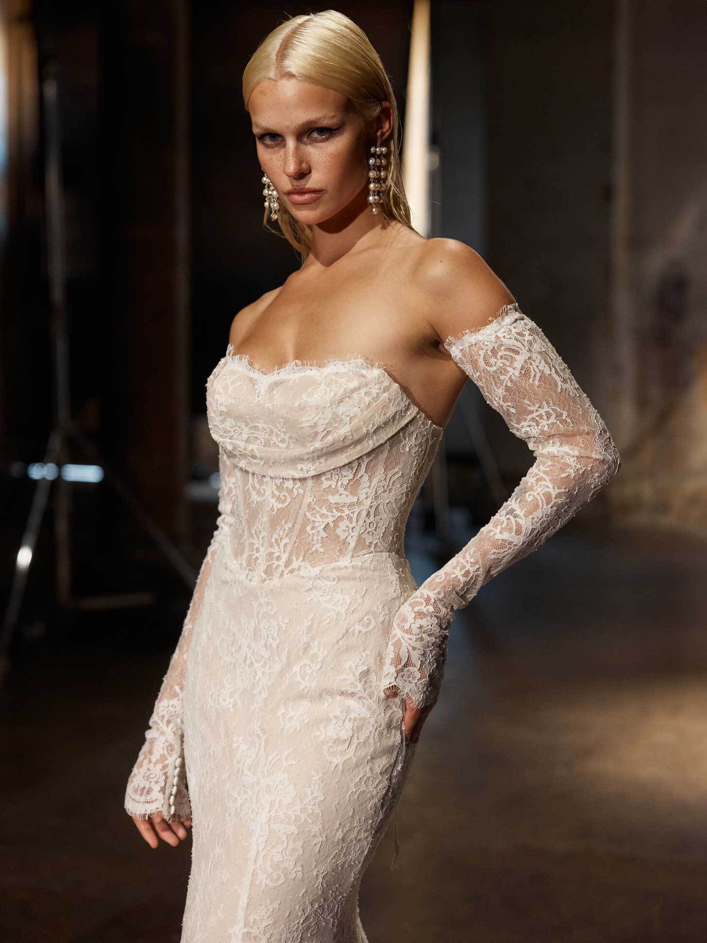 Bride in an off-the-shoulder lace gown with long sleeves, posing confidently in an industrial-style setting, her blonde hair sleek and styled, and wearing statement earrings.