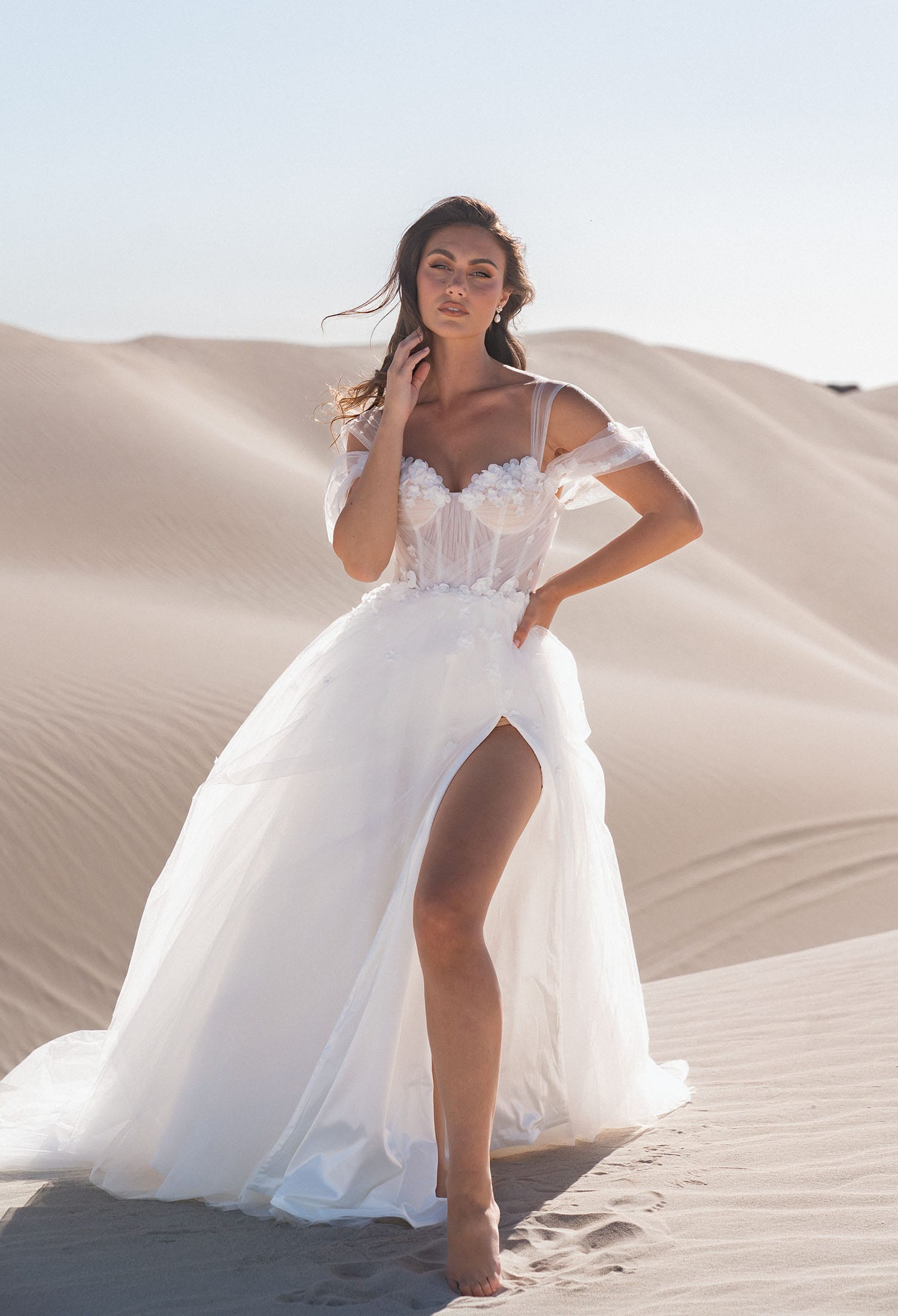 The image shows a woman wearing a stunning white wedding gown with a romantic and modern design. The dress features an off-the-shoulder neckline with delicate, sheer sleeves that drape gracefully over her arms. The bodice is fitted and adorned with intricate floral lace detailing, enhancing the romantic and feminine look. The skirt is flowing and airy, made of light, sheer fabric that adds movement and grace. A high slit on one side adds a touch of contemporary flair and reveals the woman's leg. She is wearing strappy high-heeled sandals that complement the elegant design of the dress. The woman's hair is styled in loose waves, complementing the elegant and relaxed design of the gown. The overall effect is one of ethereal beauty and sophistication, perfect for a modern fairy-tale wedding.