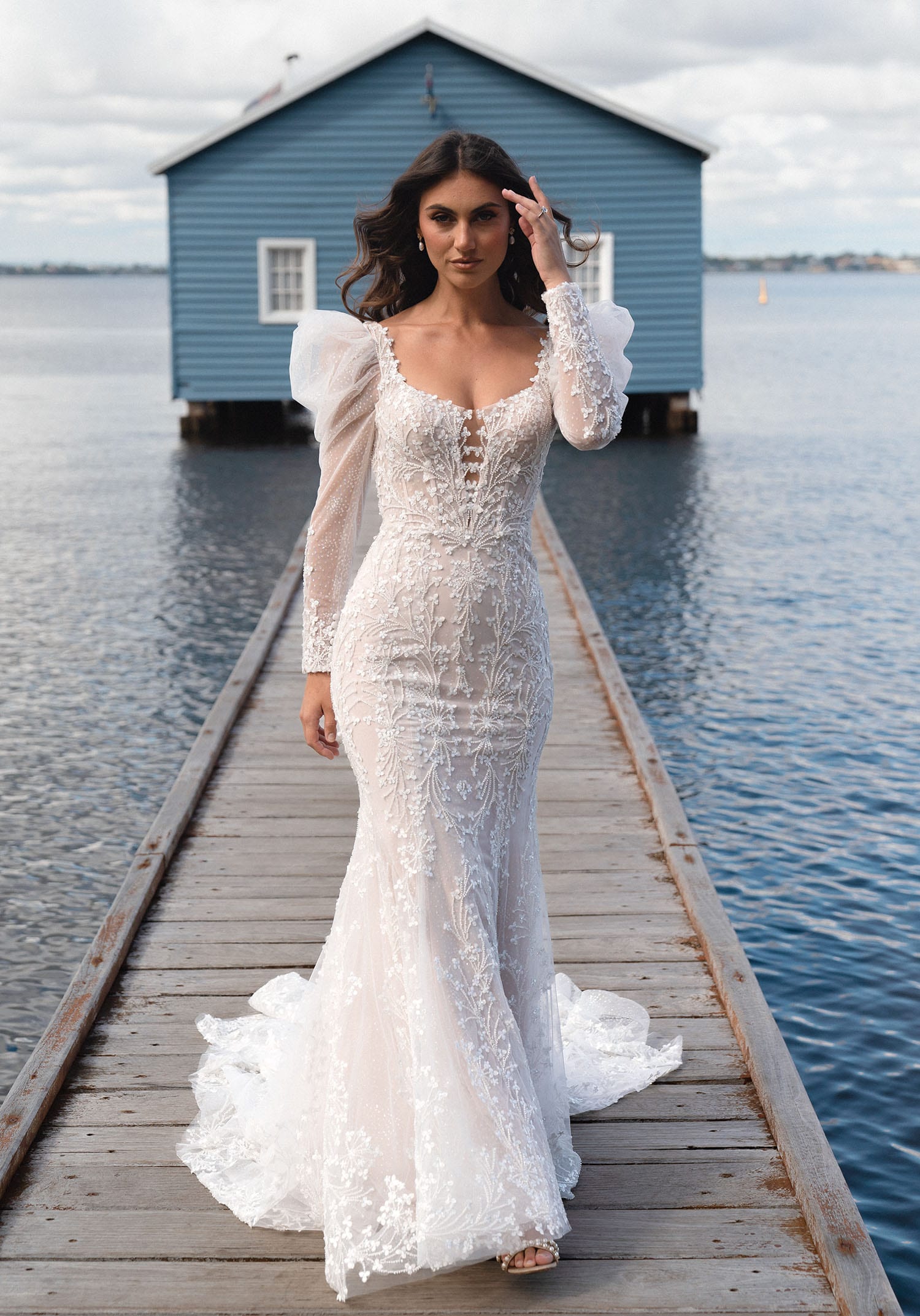 The image shows a woman wearing a stunning and elegant white wedding gown. The dress features a deep V-neckline with delicate, sheer long sleeves that have a puffed shoulder detail, adding a touch of vintage charm to the modern design. The bodice is intricately adorned with lace and beadwork, creating a sophisticated and romantic look. The gown follows a slim, fitted silhouette that hugs the body and flares out slightly at the bottom into a small, graceful train. The lace detailing continues throughout the length of the dress, adding texture and visual interest. The woman's hair is styled in loose waves, complementing the elegant and timeless design of the gown. The overall effect is one of refined beauty, combining modern elegance with a touch of classic romance, perfect for a stylish and contemporary wedding.