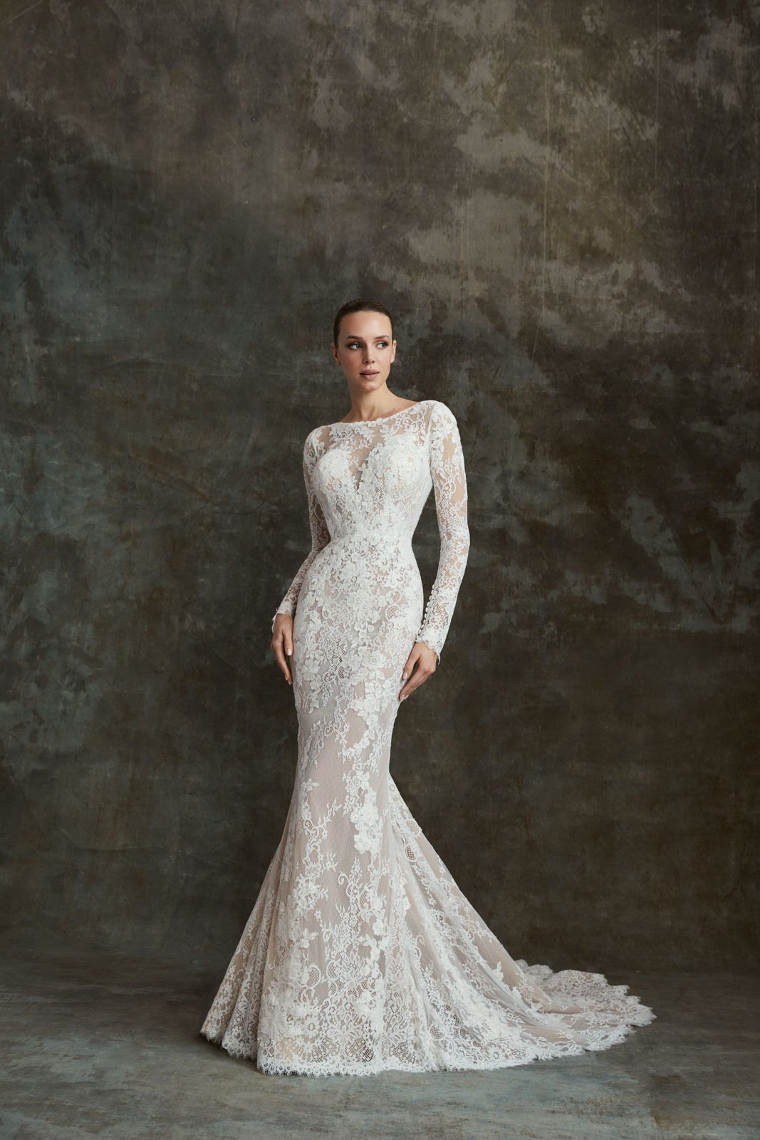 The image showcases the Shirley wedding gown, a full-length lace dress featuring a figure-hugging silhouette that flares out into a delicate, sweeping train. The gown is made entirely of intricate lace, with a high bateau neckline and long, fitted sleeves that exude timeless elegance. The lace pattern, composed of floral motifs, covers the dress from top to bottom, creating a luxurious texture and a sophisticated, classic look. The train extends gracefully behind the bride, adding a touch of drama to the overall design. The neutral backdrop highlights the gown's details, making it the focal point of the image. This dress is perfect for a bride who seeks a blend of tradition and refined style.