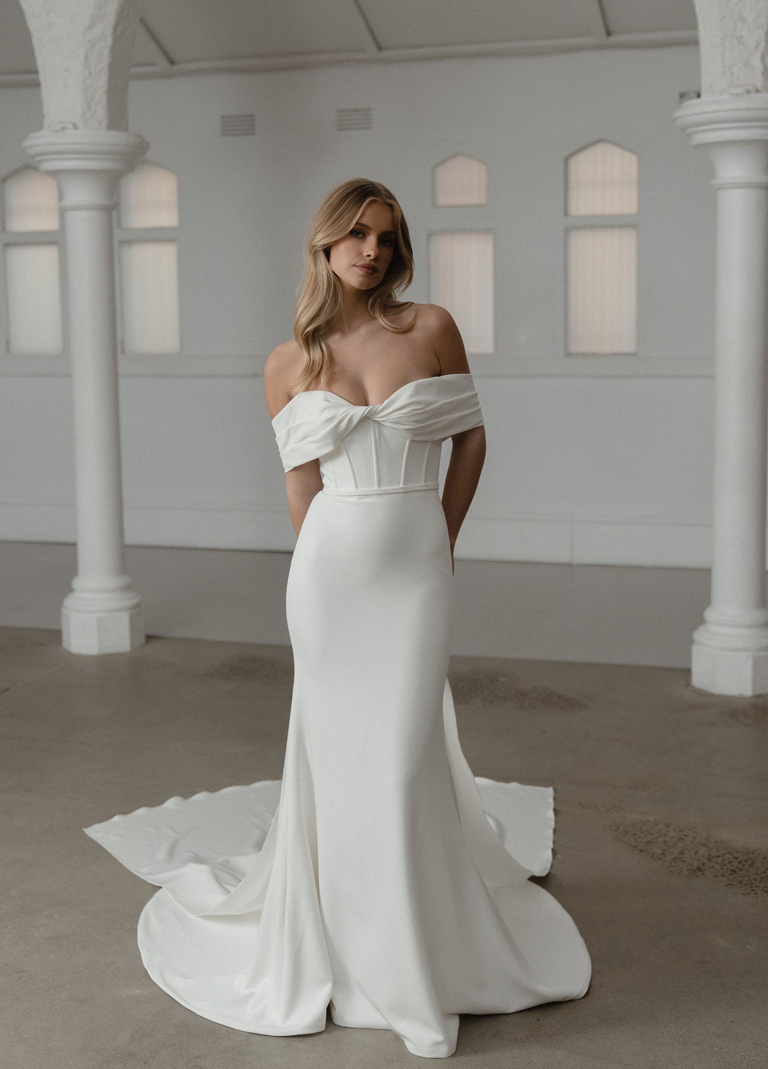 The image shows a woman wearing an elegant white wedding gown. The dress features an off-the-shoulder design with draped sleeves that create a romantic and sophisticated look. The bodice is fitted and structured, highlighting the woman's figure, with a slight V-neckline adding a touch of elegance. The skirt is sleek and flows gracefully to the floor, extending into a subtle train behind her. The fabric appears smooth and luxurious, contributing to the gown's refined and timeless appearance. The woman's hair is styled in loose waves, cascading over her shoulders, complementing the chic and elegant design of the dress. The overall effect is one of understated elegance and modern sophistication, perfect for a contemporary wedding.