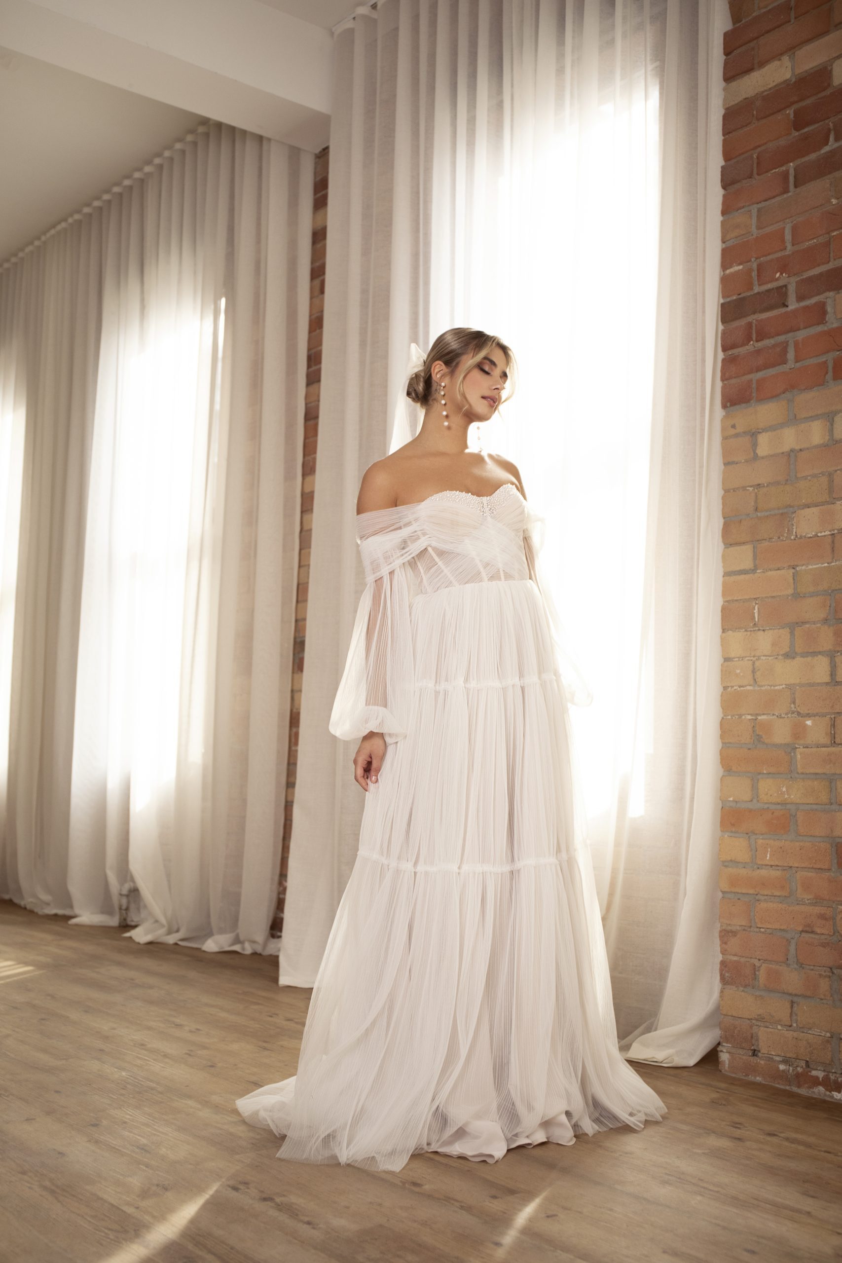 A bride stands gracefully in a Sarah Alouache Bridal gown, bathed in the warm glow of natural light streaming through sheer curtains. The dress, with its off-the-shoulder design and voluminous, layered skirt, exudes an ethereal, romantic charm. The soft, flowing fabric of the gown complements the rustic brick wall backdrop, creating a beautiful contrast between the delicate bridal look and the textured surroundings. Her hair is styled in a loose, elegant bun, adorned with a subtle ribbon, while statement pearl earrings add a touch of sophistication to the overall ensemble.