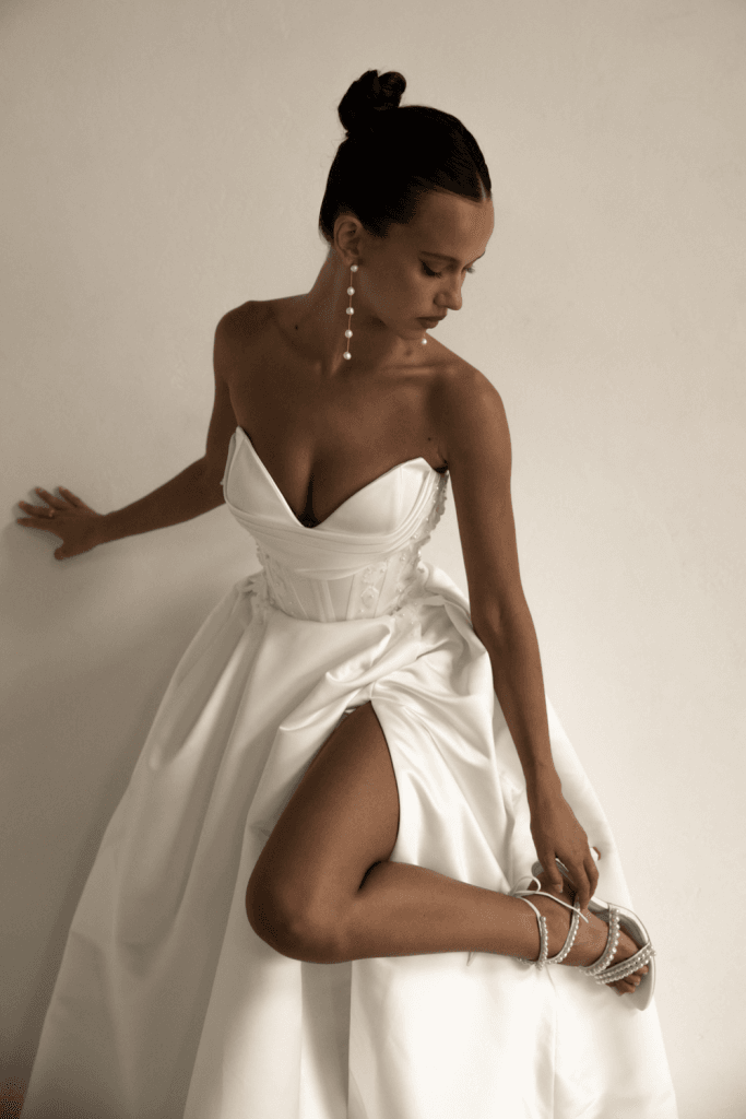 The image shows a woman wearing a strapless white wedding dress with a sweetheart neckline and a dramatic thigh-high slit on the left side.  showing a close up action shot of the front of the bodice. The dress has a voluminous skirt that falls gracefully to the floor, creating a flowing train. The woman is posing with her right hand resting on a plain wall and her left hand on her hip. She is wearing white strappy high-heeled sandals that are visible through the slit. The setting is simple and minimalistic, with a light-colored wall and a neutral floor, focusing all attention on the woman and her dress.