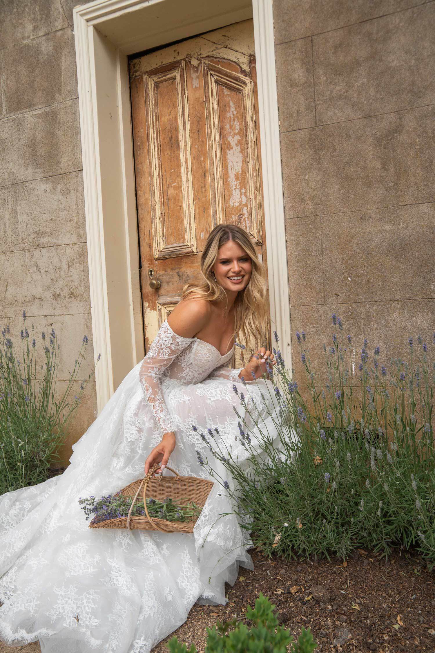 Bride in an off-the-shoulder lace gown, kneeling in a garden with lavender plants, holding a wicker basket, and smiling in front of a weathered wooden door.